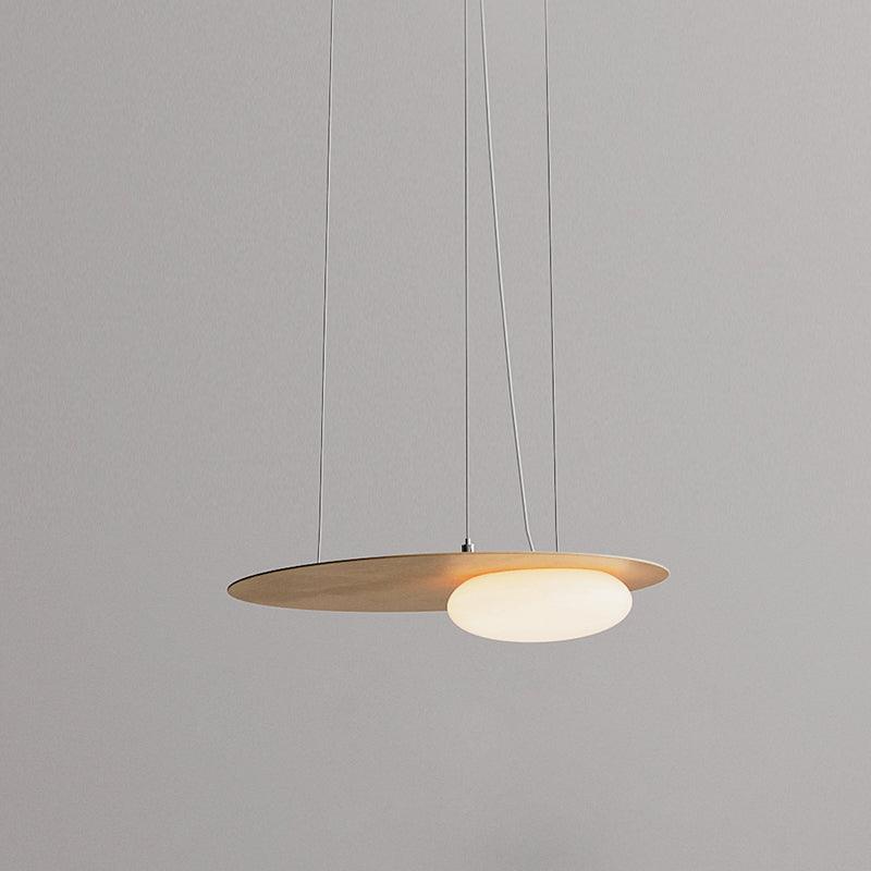 Gold disc pendant against grey, with offset glowing white diffuser, fine support cables and a pale curved power cord.