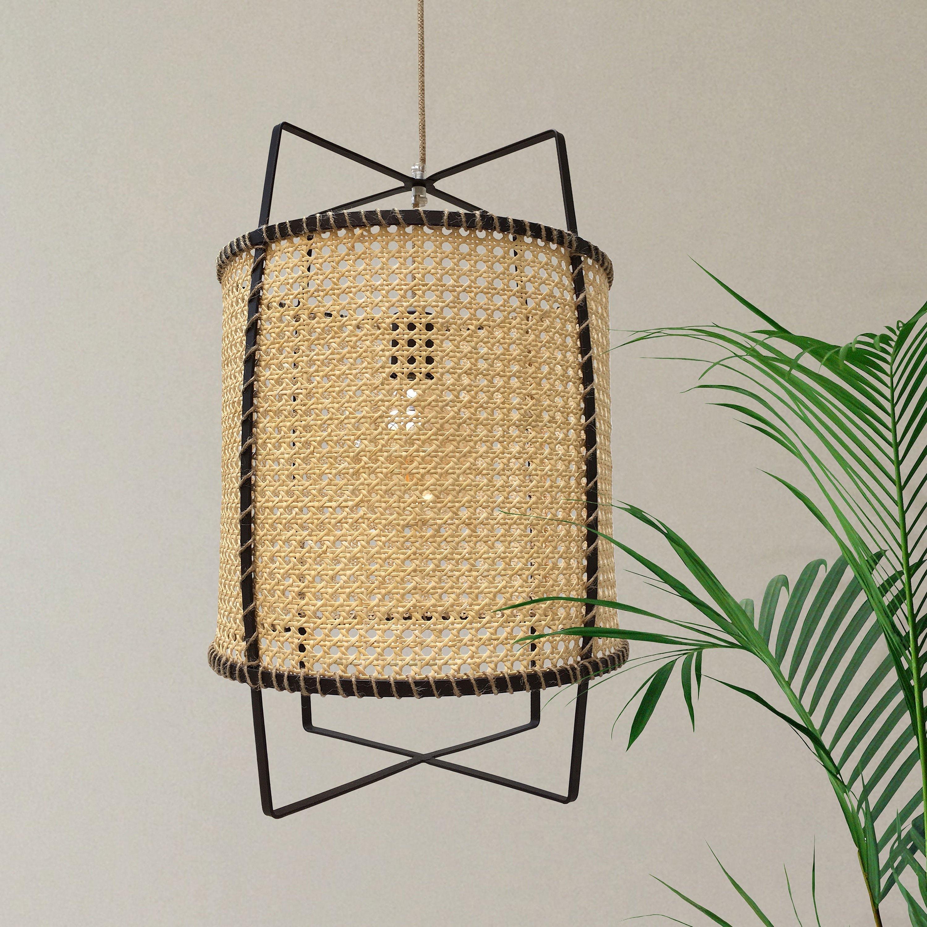Unlit natural cane pendant with dark geometric frame beside palm leaves on a pale wall