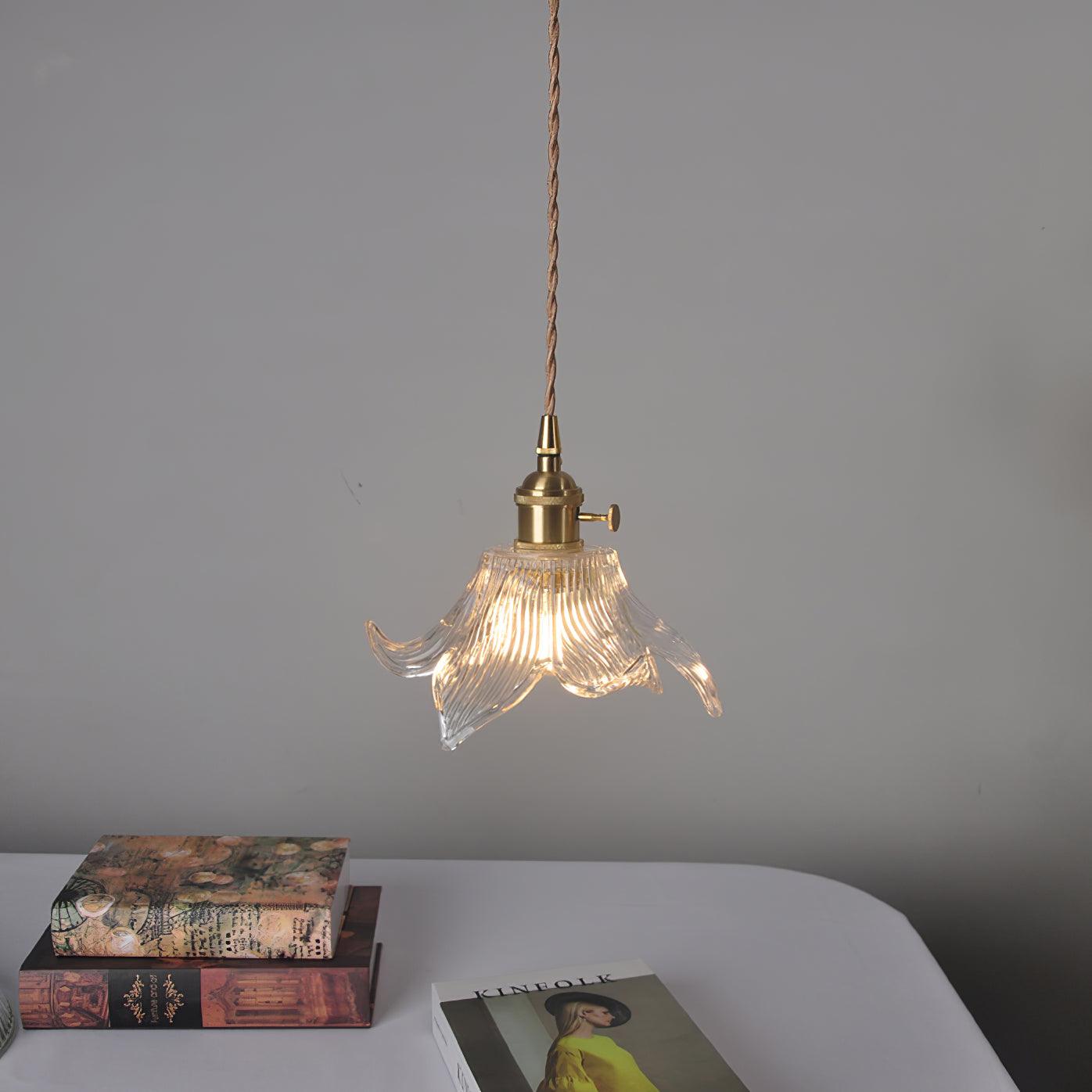 Wider view of the lit flower glass pendant on its twisted cord above a white rounded table with books.