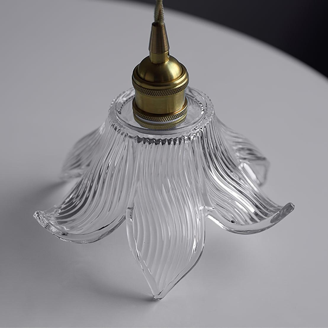 Unlit upper angled view of the knurled brass-toned holder, clear ribbed petal shade and tan cord above a white table.