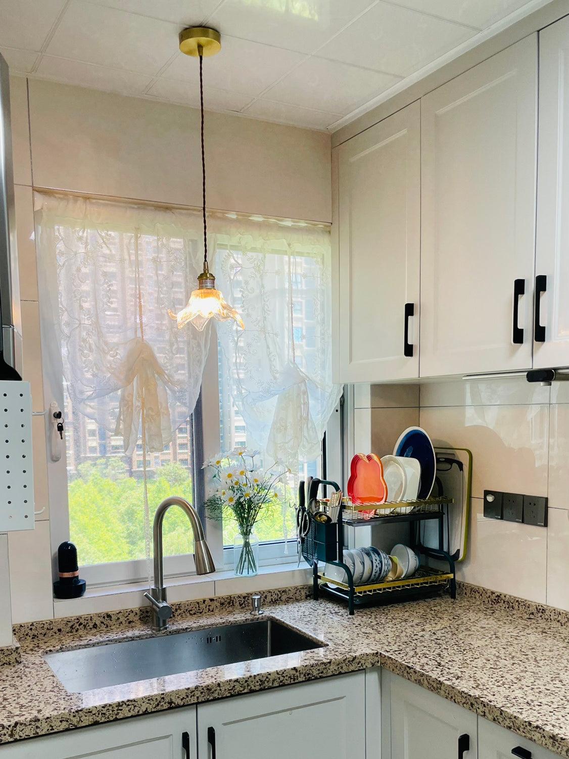 Single lit petal pendant with round ceiling canopy above a kitchen sink, lace curtains, speckled counter, white cabinets and plate rack.