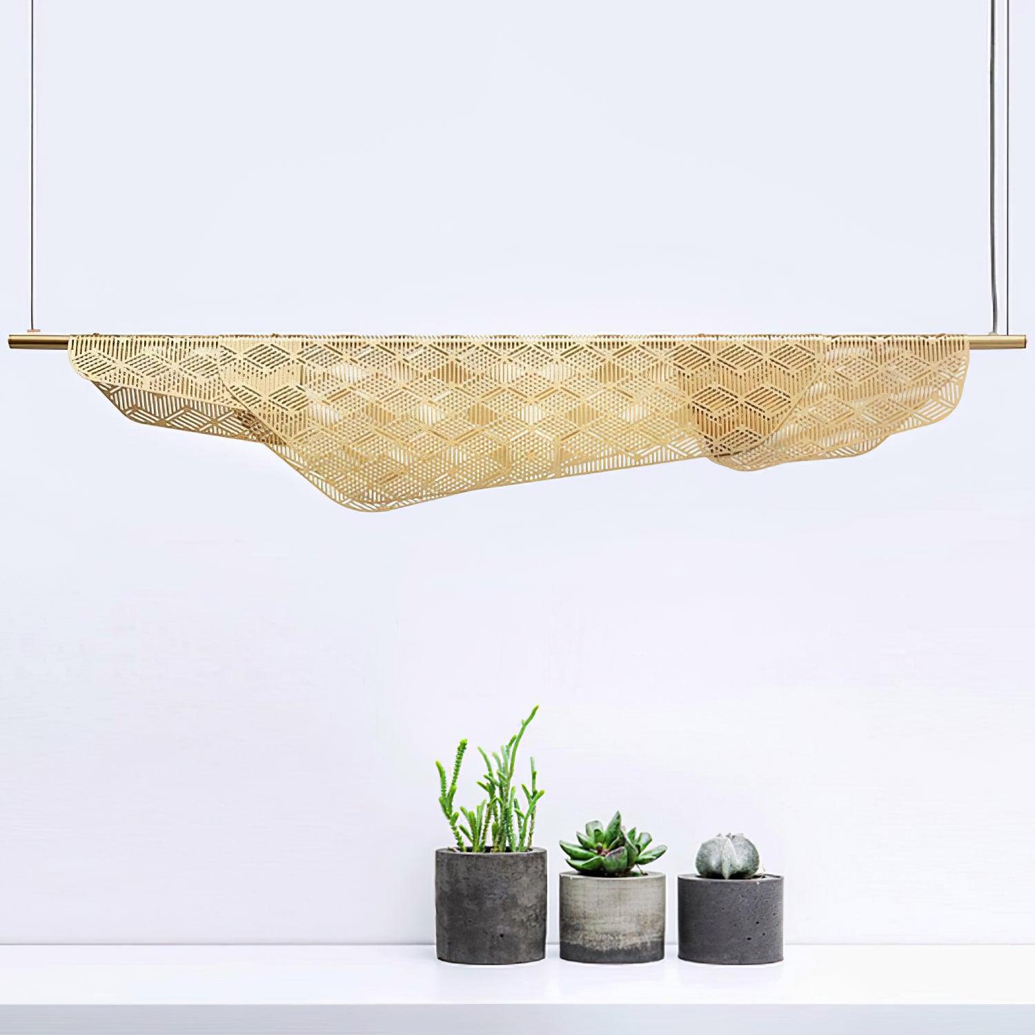 Long unlit gold perforated pendant above three small potted succulents on a white surface, showing the draped lower edge and fine suspension lines.