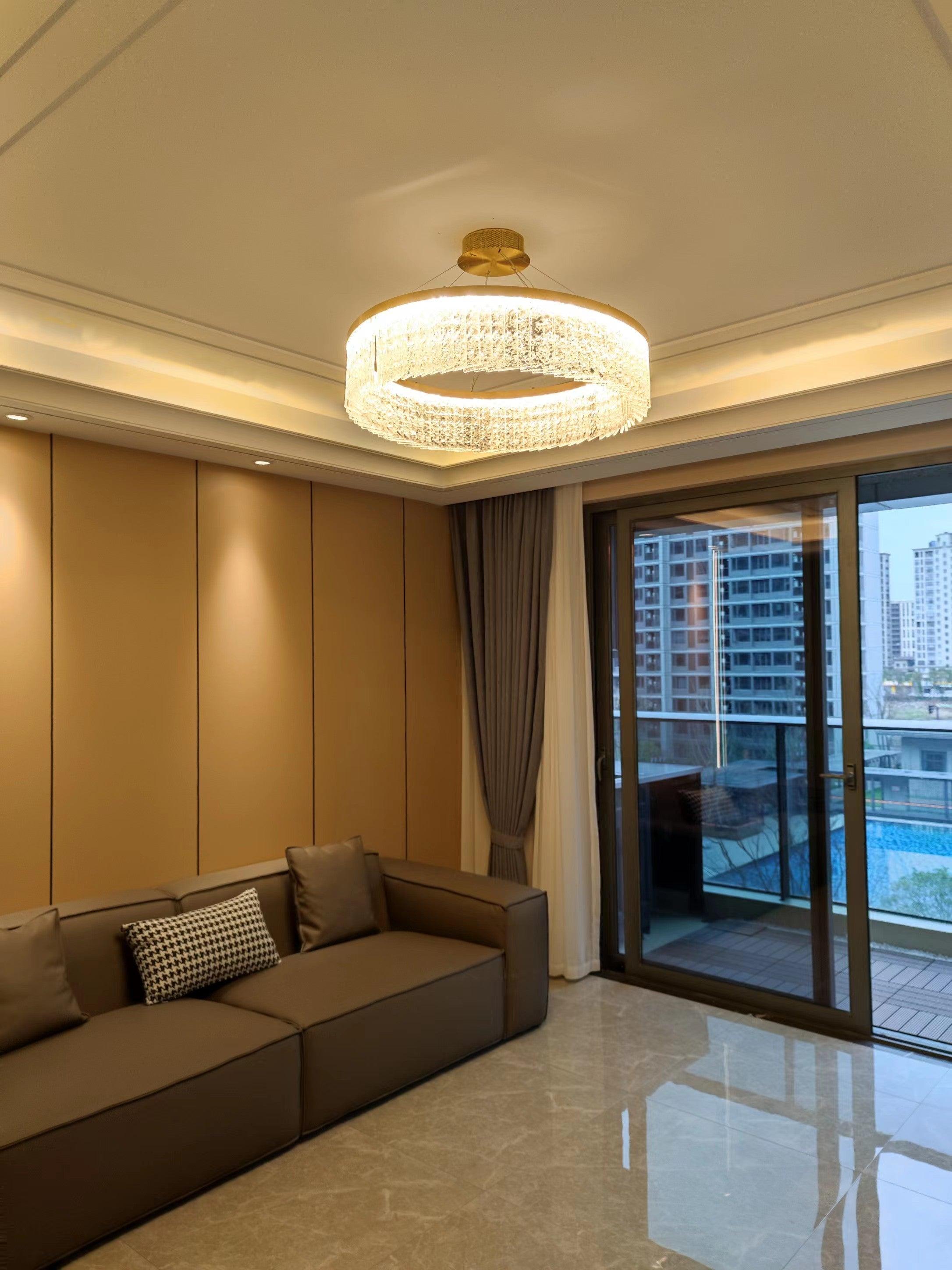 Short-hanging illuminated ring above brown sofa near balcony glazing and city view, with warm ceiling cove lights