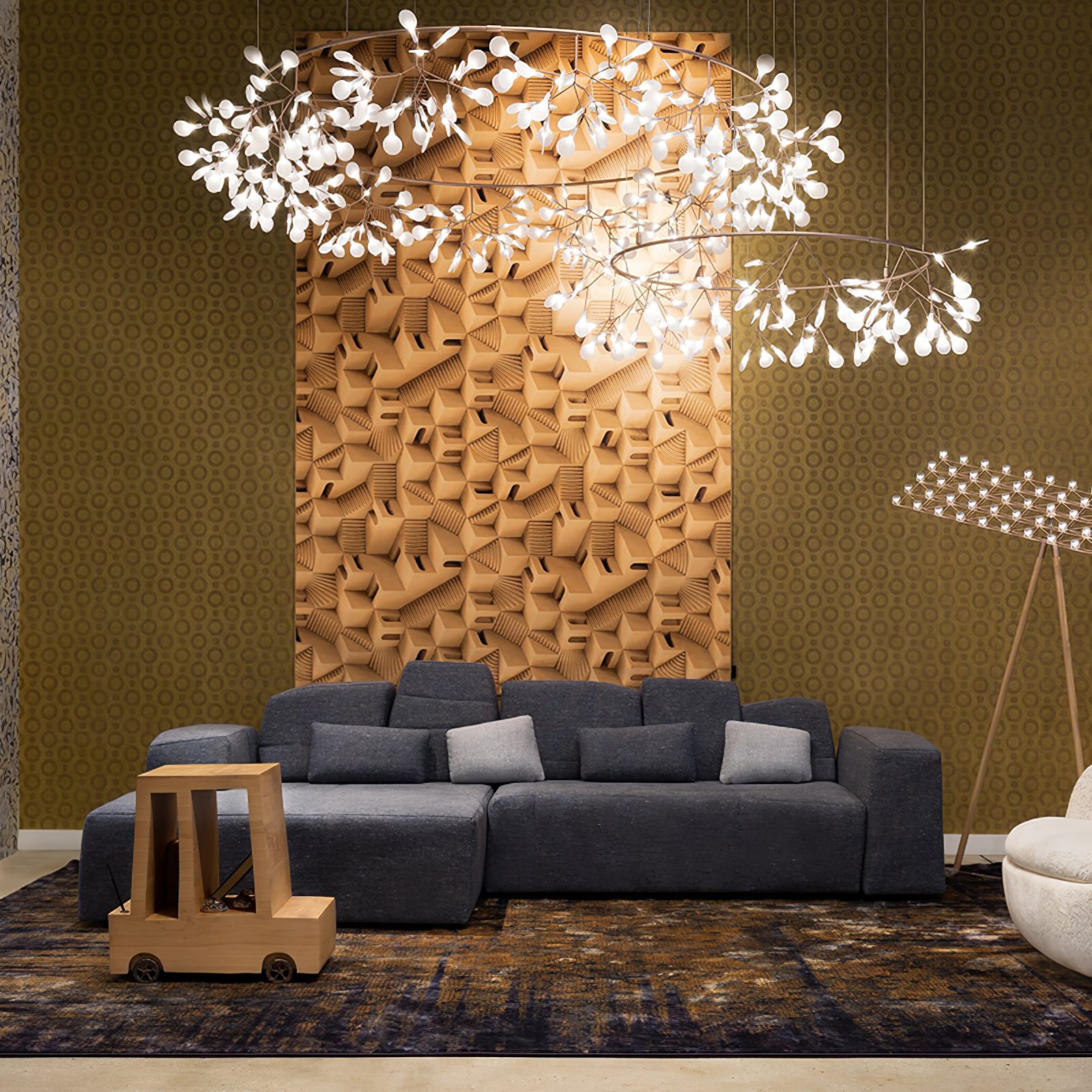 Two warm metallic leaf ring chandeliers above a dark sectional sofa against a mustard geometric wall