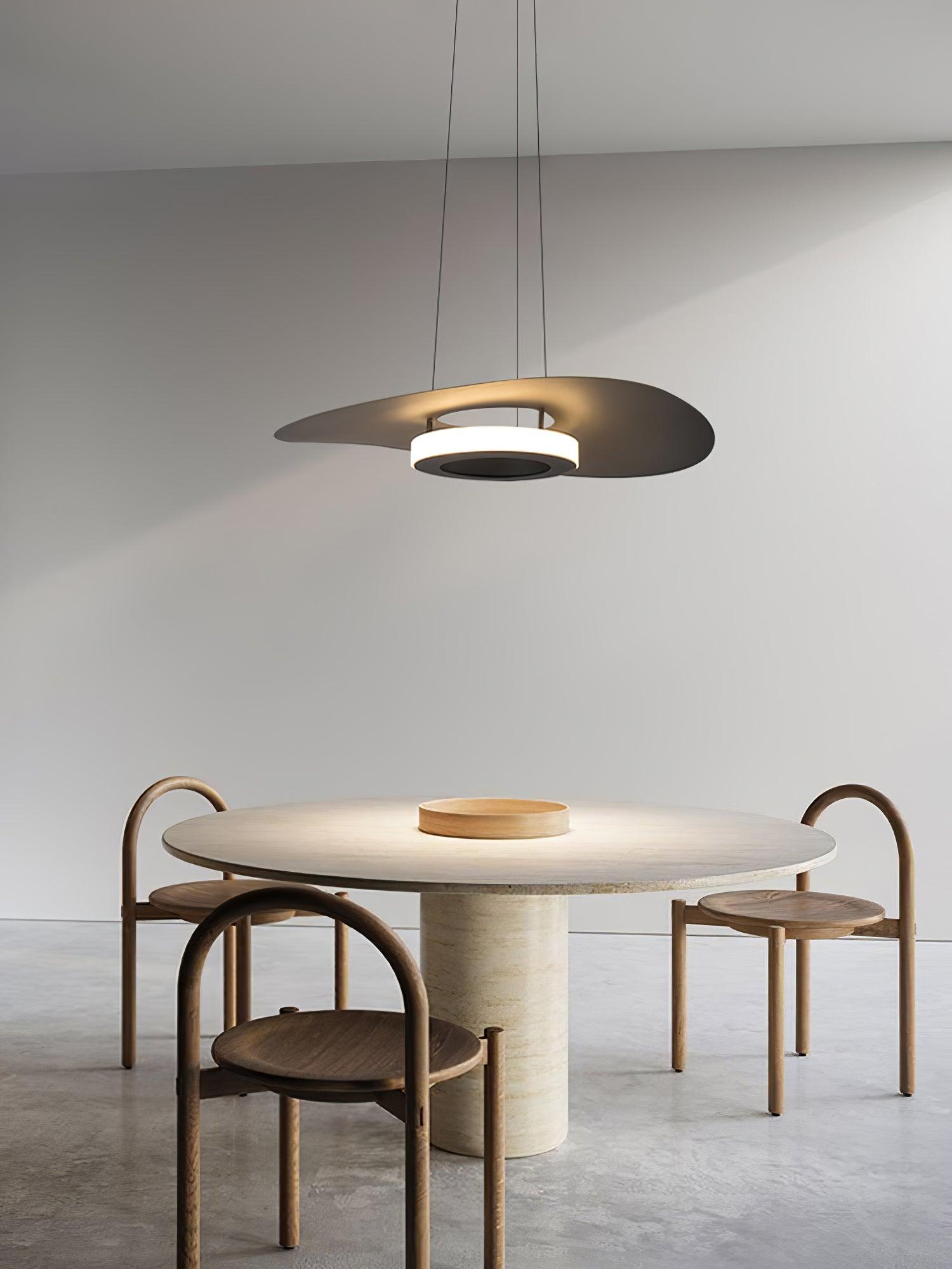 Dark curved disc pendant above round stone pedestal table with shallow wooden tray and arched wooden chairs