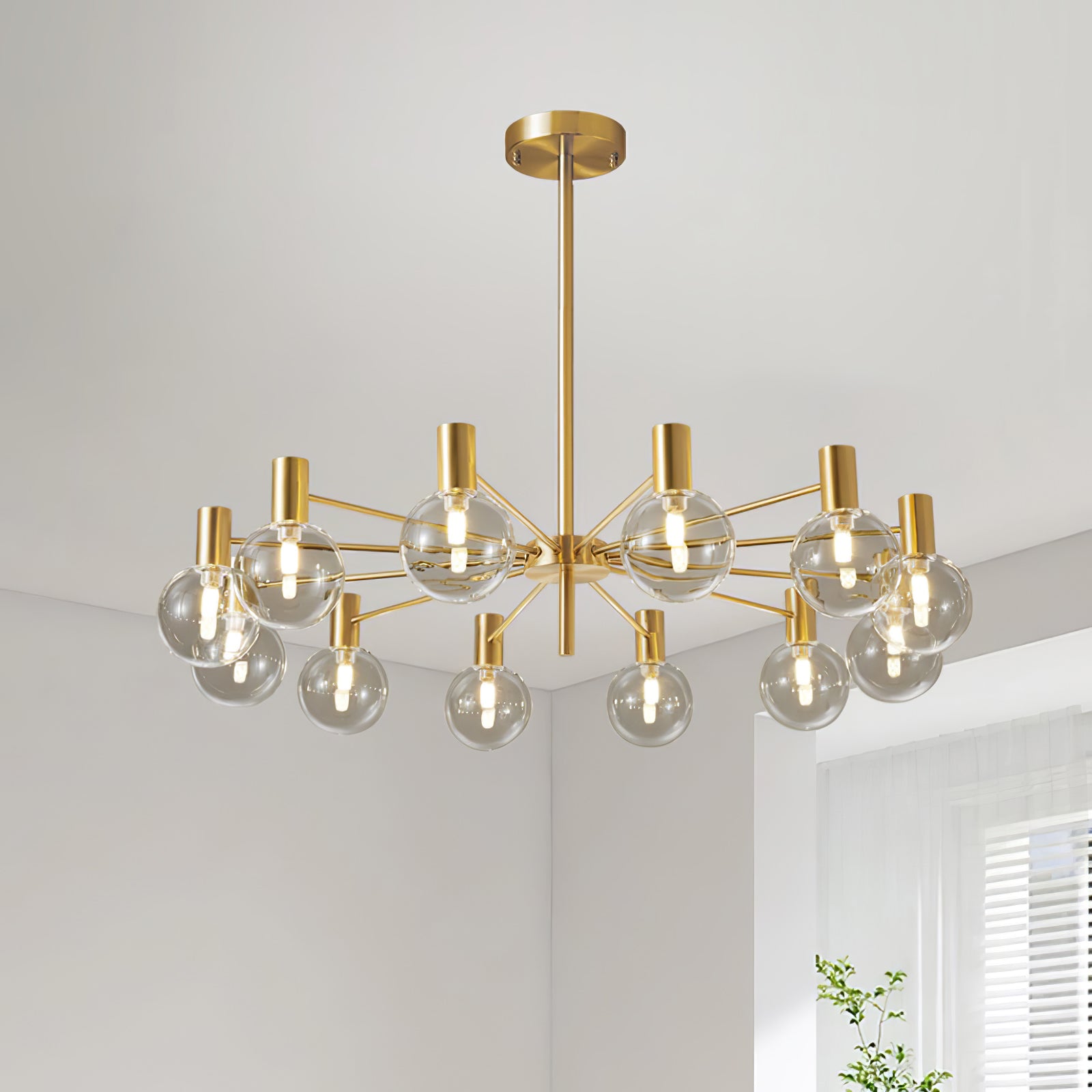 Twelve-clear-globe chandelier with gold-tone arms, rod and canopy in a pale room beside a white blind and green plant.