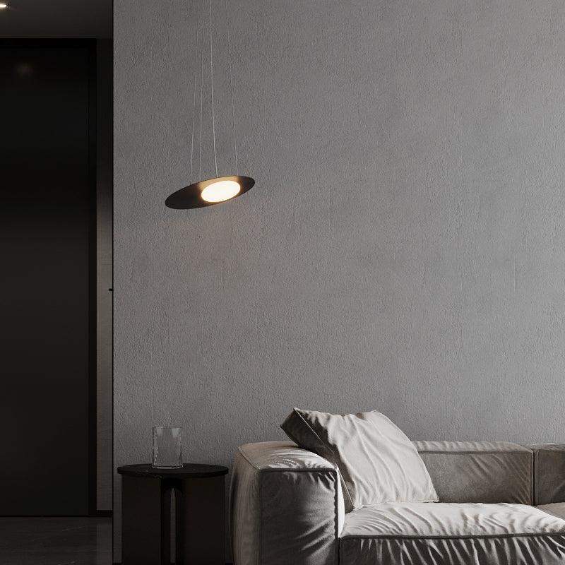 Black disc pendant beside a grey sofa, above a round black side table and clear vase, against textured grey wall.