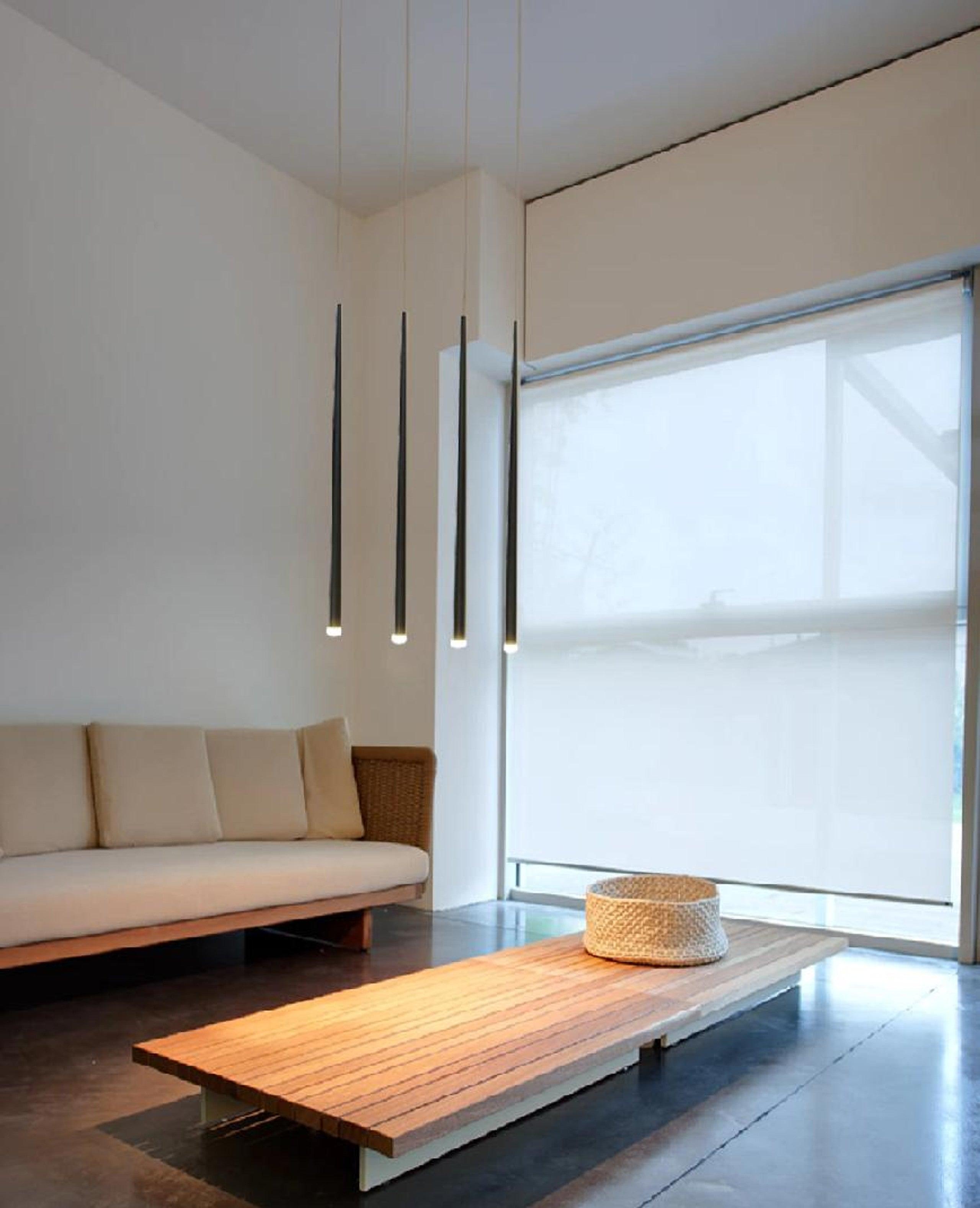 Four glowing black tube pendants near a window above a cream sofa and slatted wooden coffee table