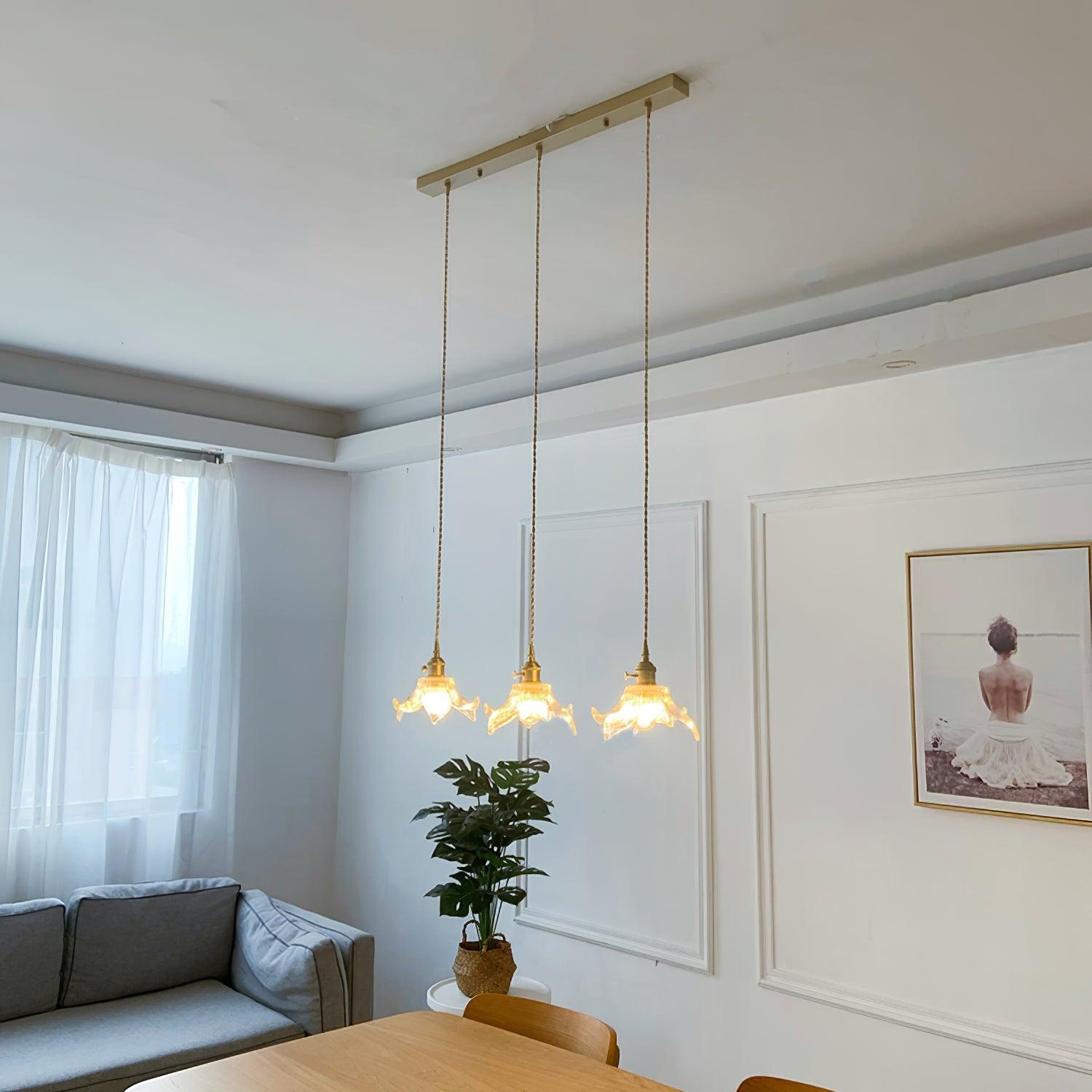 Three lit petal pendants on a shared rectangular brass-toned ceiling bar above a timber dining table beside a grey sofa and monstera.