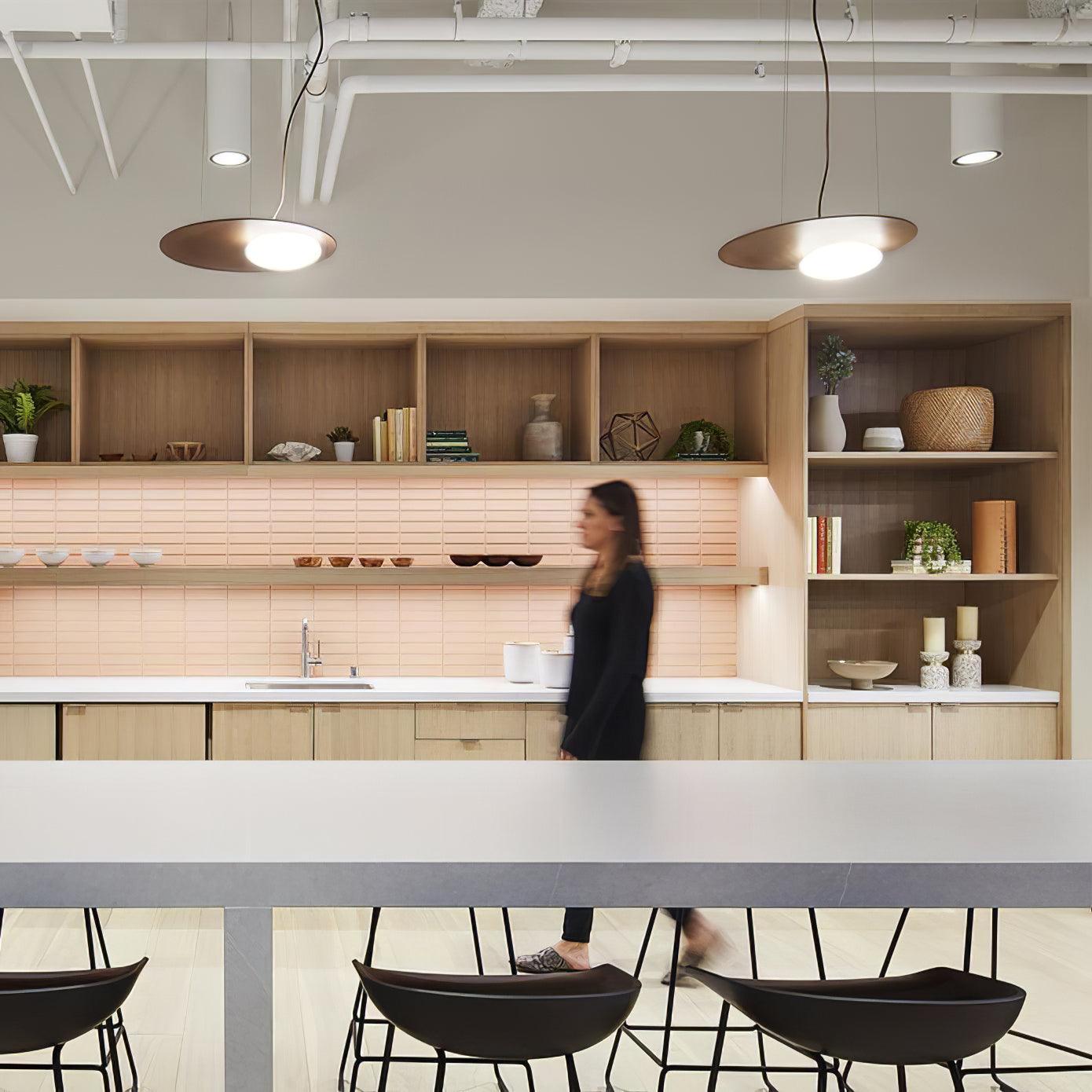 Two gold-tone disc pendants in a kitchen with exposed ceiling pipes, wood shelving and pink tiles; pale island and black stools below.
