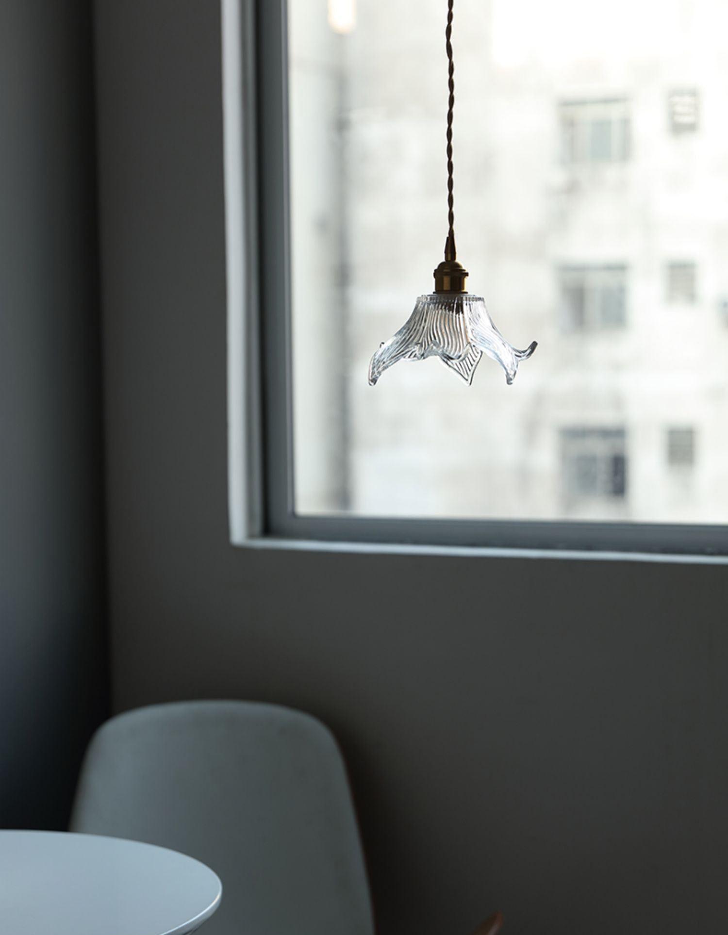 Single unlit clear flower glass pendant against a large window, above a white round table and pale upholstered chair.