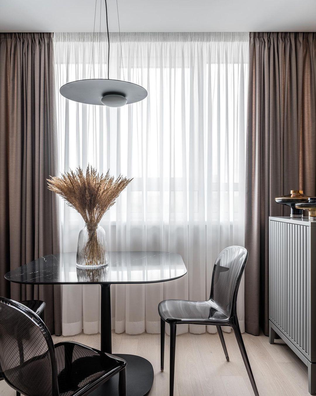 Black pendant over glossy table and dried-grass vase by sheer and brown curtains, with dark transparent chairs and ribbed cabinet.