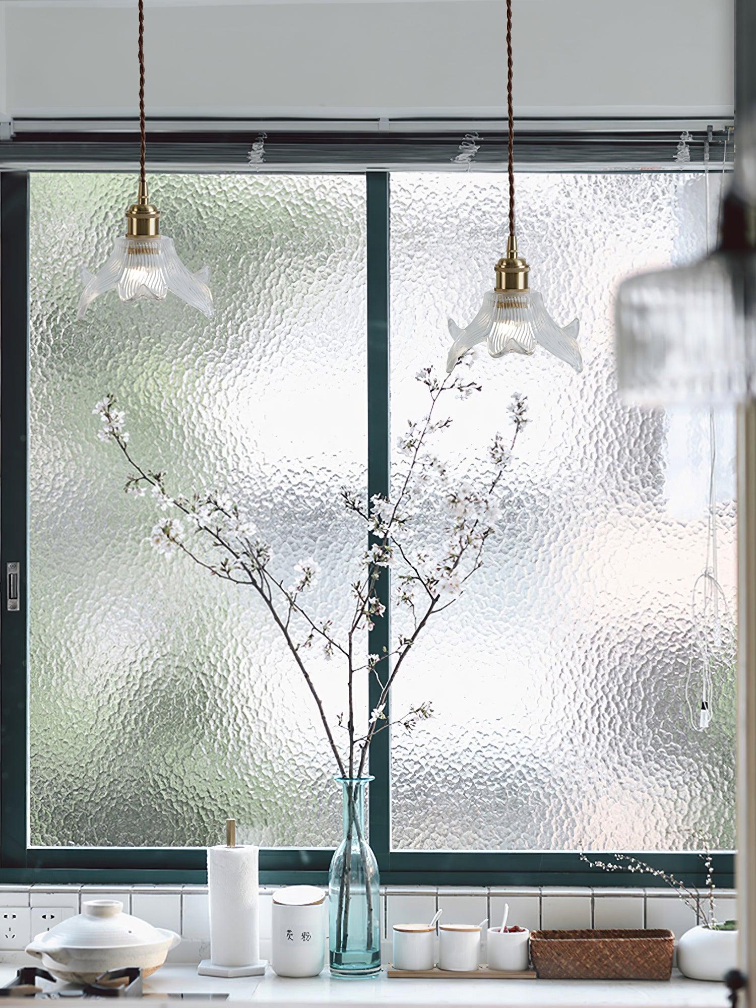 Two lit petal glass pendants before a textured window above white kitchen pots and tall blue glass vase with flowering branches.