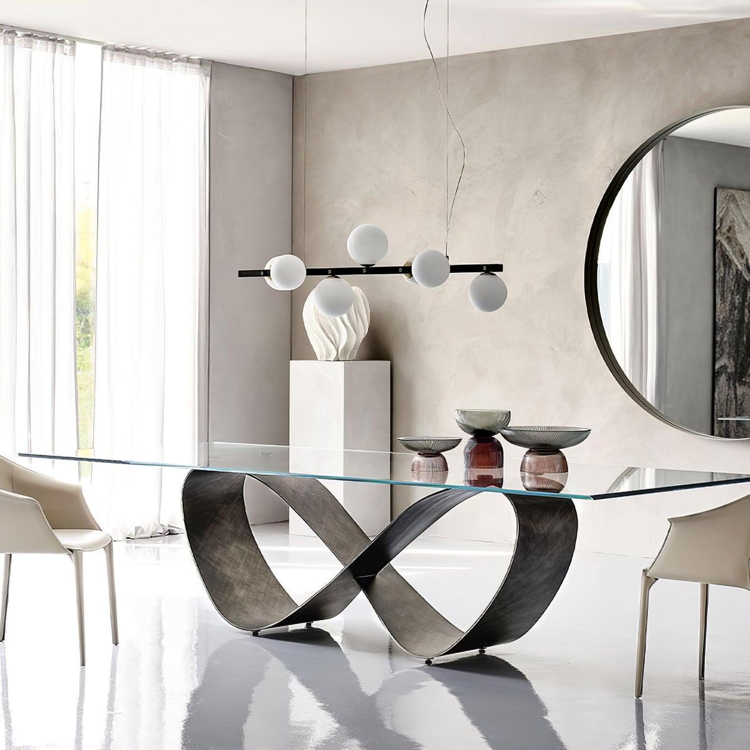 Globe pendant above clear table and looped metal base, with white chairs, round wall mirror, sculptural ornament and coloured vases.