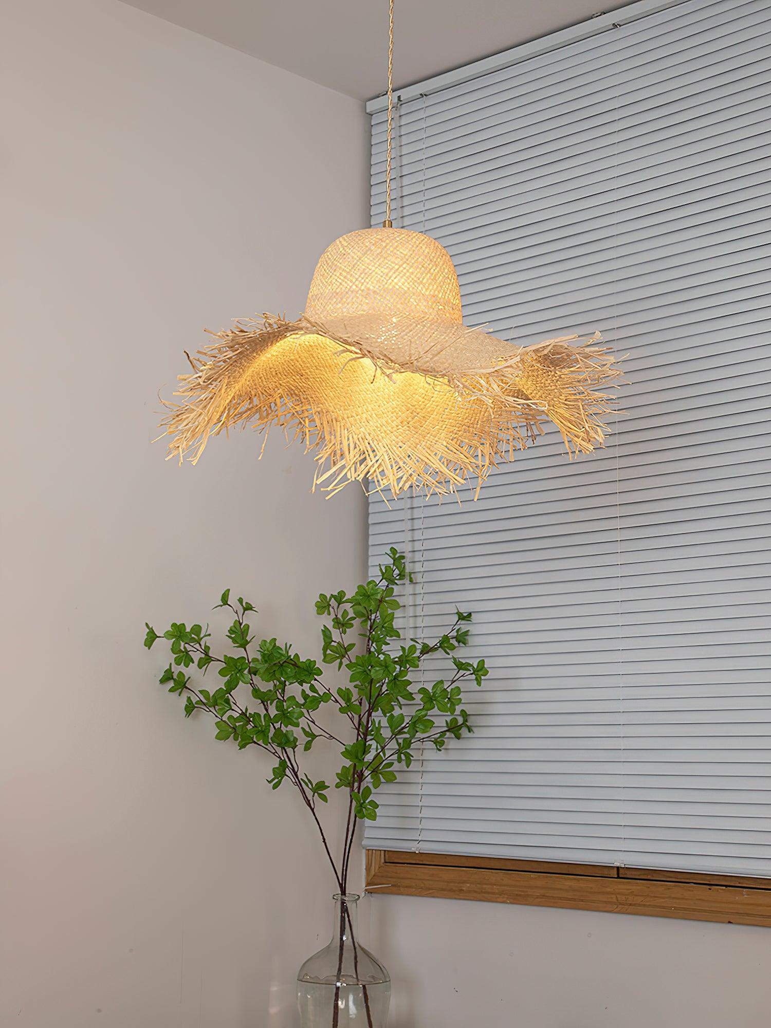 Low view of the glowing woven hat shade and fringed underside before white blinds and a tall leafy vase