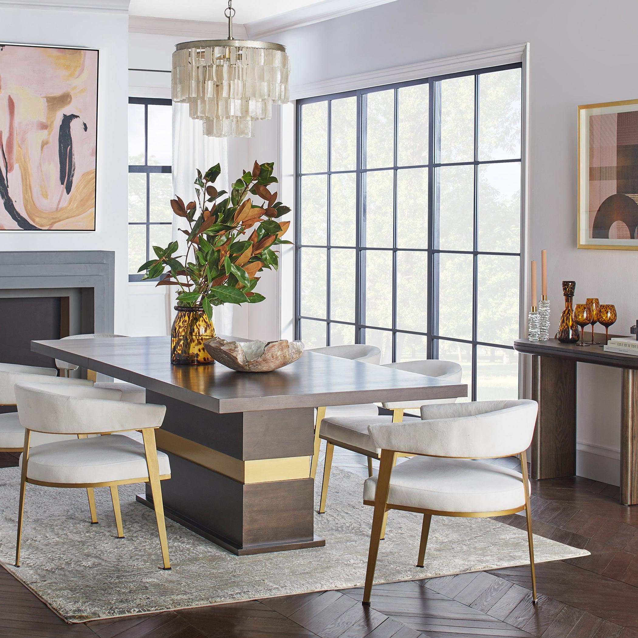 Shell pendant above a dark dining table with white upholstered chairs, black-framed windows and a leafy arrangement