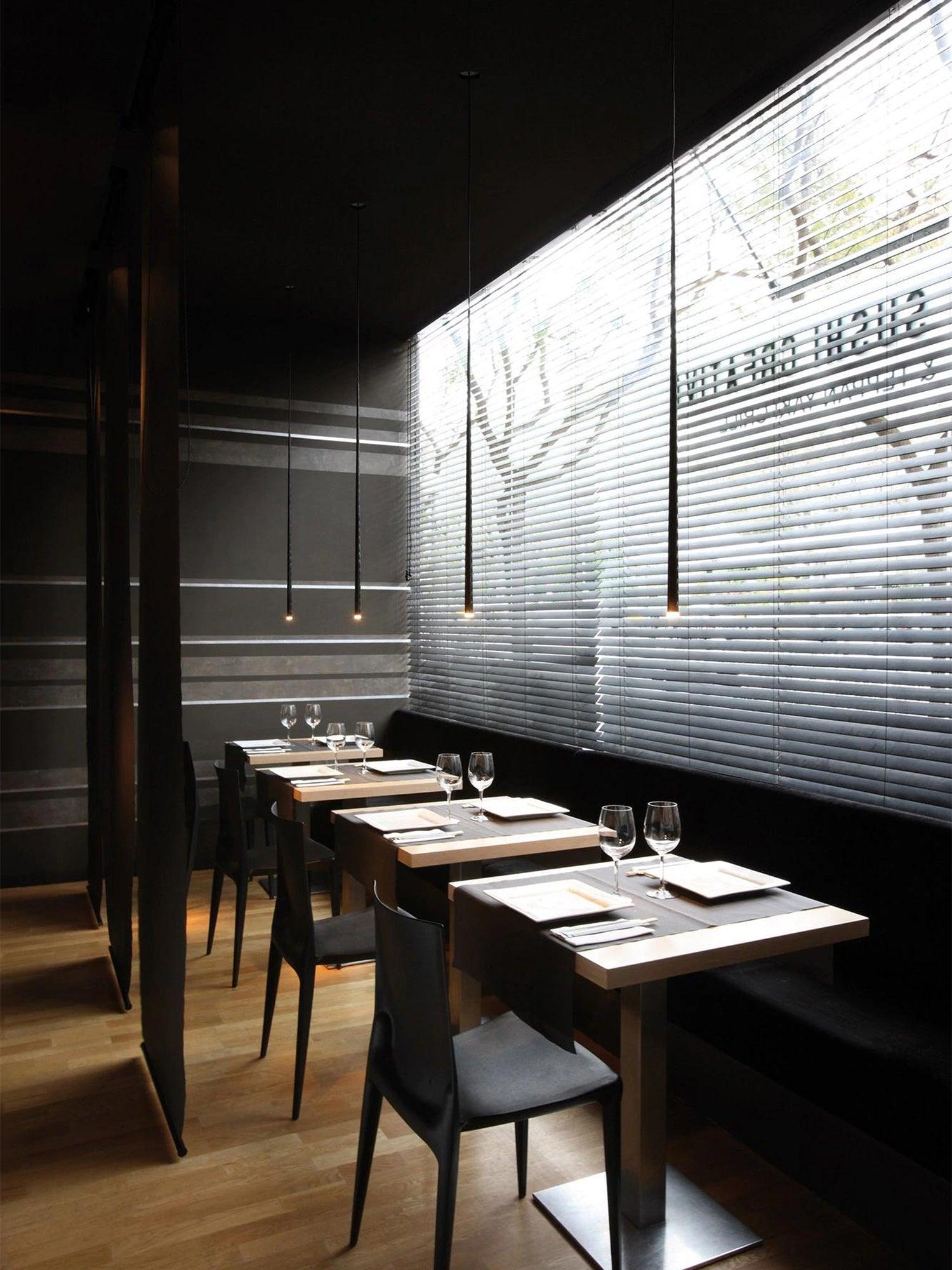 Separate black single tube pendants above small wooden restaurant tables beside window blinds