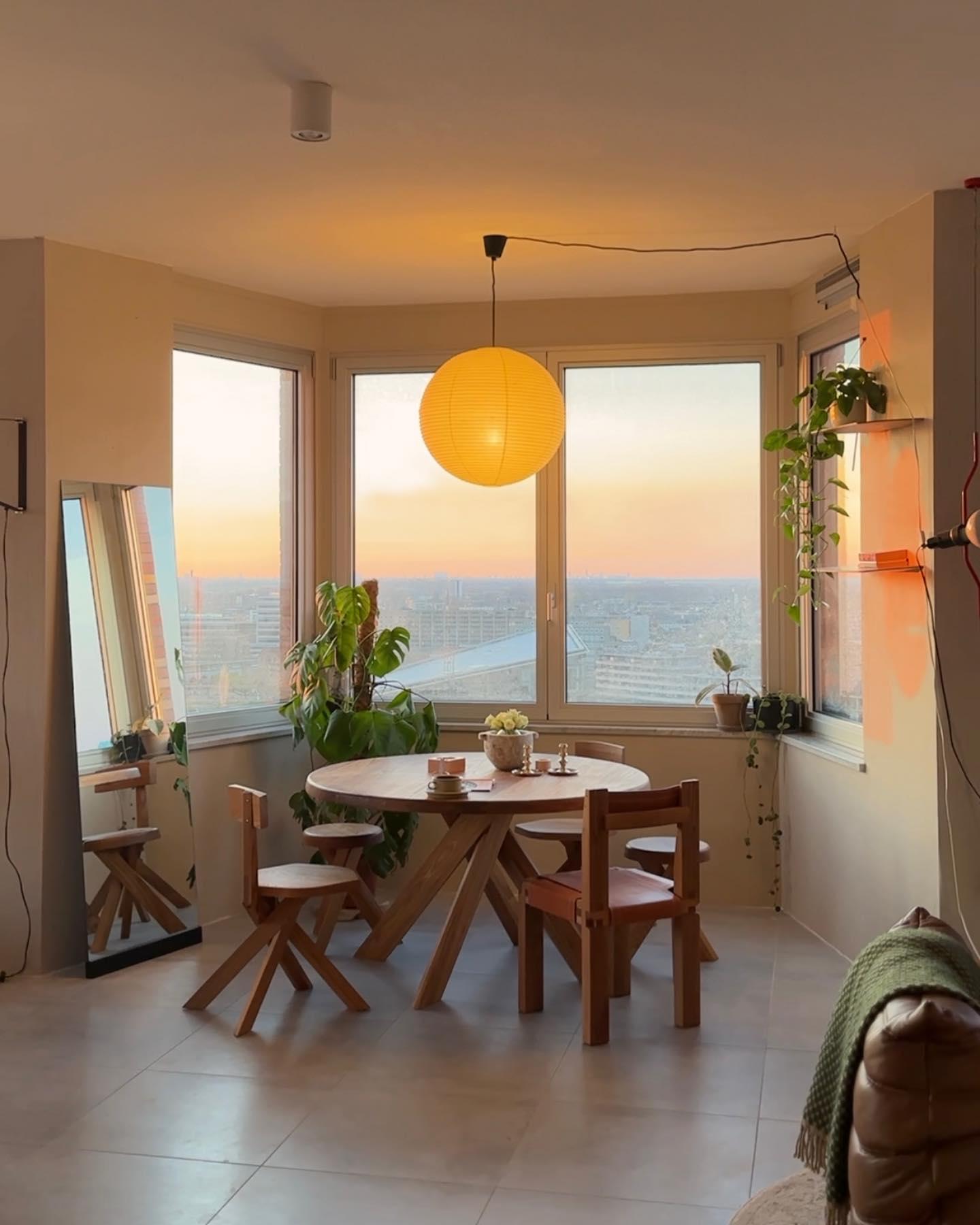 Amber-looking round lantern above wood dining table by bay windows, plants and leaning mirror