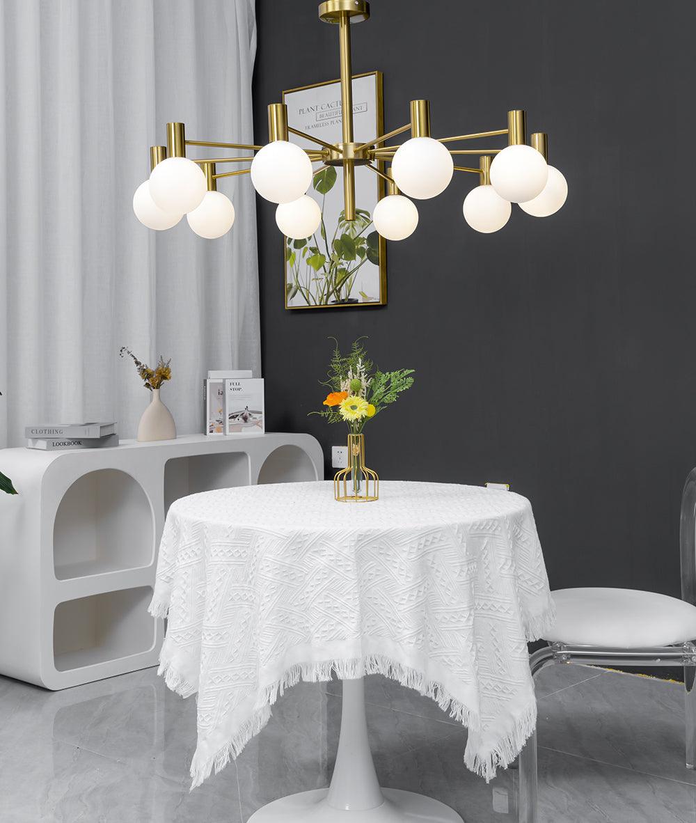 Lit white-globe chandelier above a round table with a fringed white cloth, beside a clear chair and white cubby console.