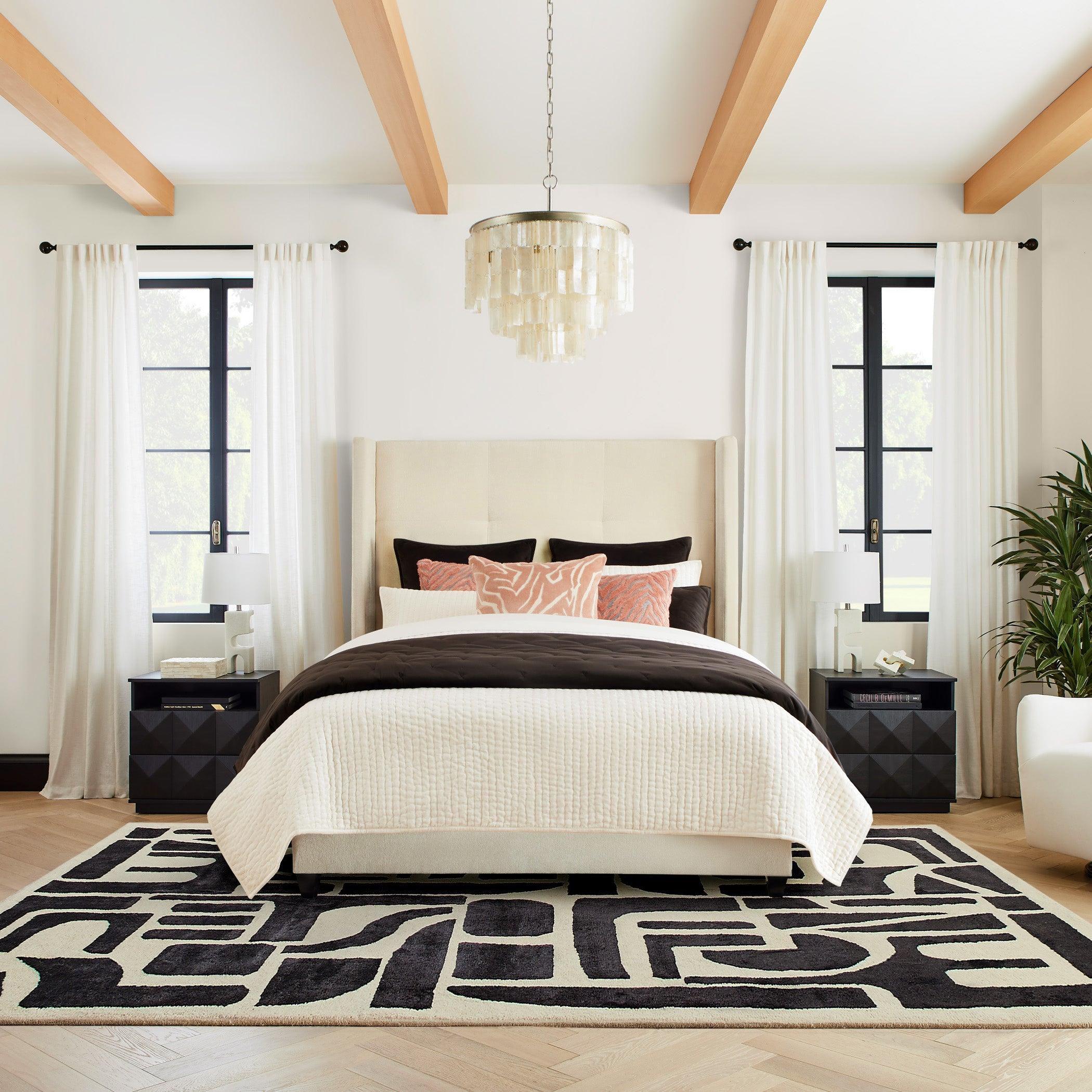 Shell pendant centred over a cream bed with coral pillows, black bedside cabinets and a black-and-white geometric rug