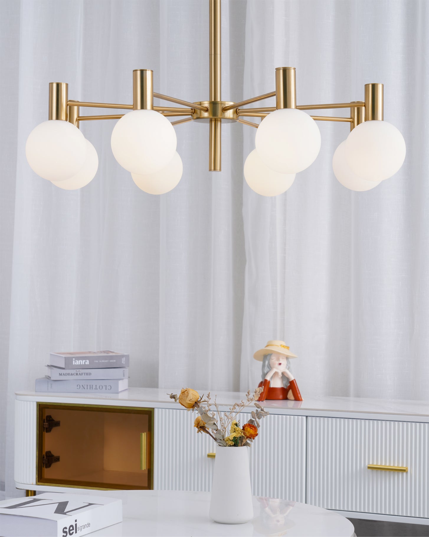 Close view of lit white globes and gold-tone arms above a cabinet with books, a figurine and dried flowers.