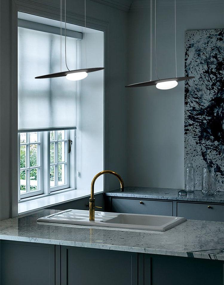 Two black glowing disc pendants over a veined sink counter with gold-tone tap, window blind, bottles and wall art.