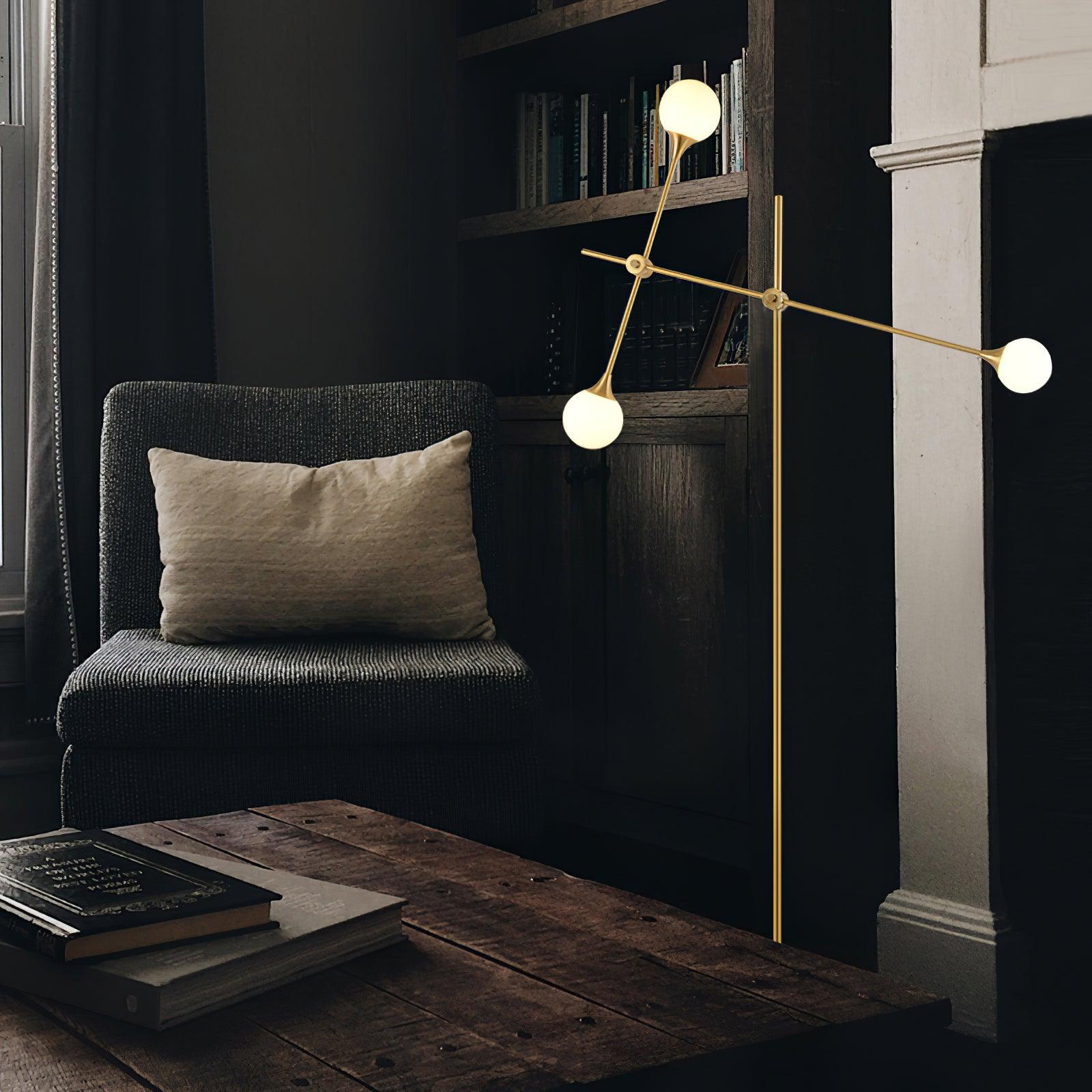 Lit gold Kemi floor lamp beside a dark armchair and bookcase, with a wooden table in the foreground