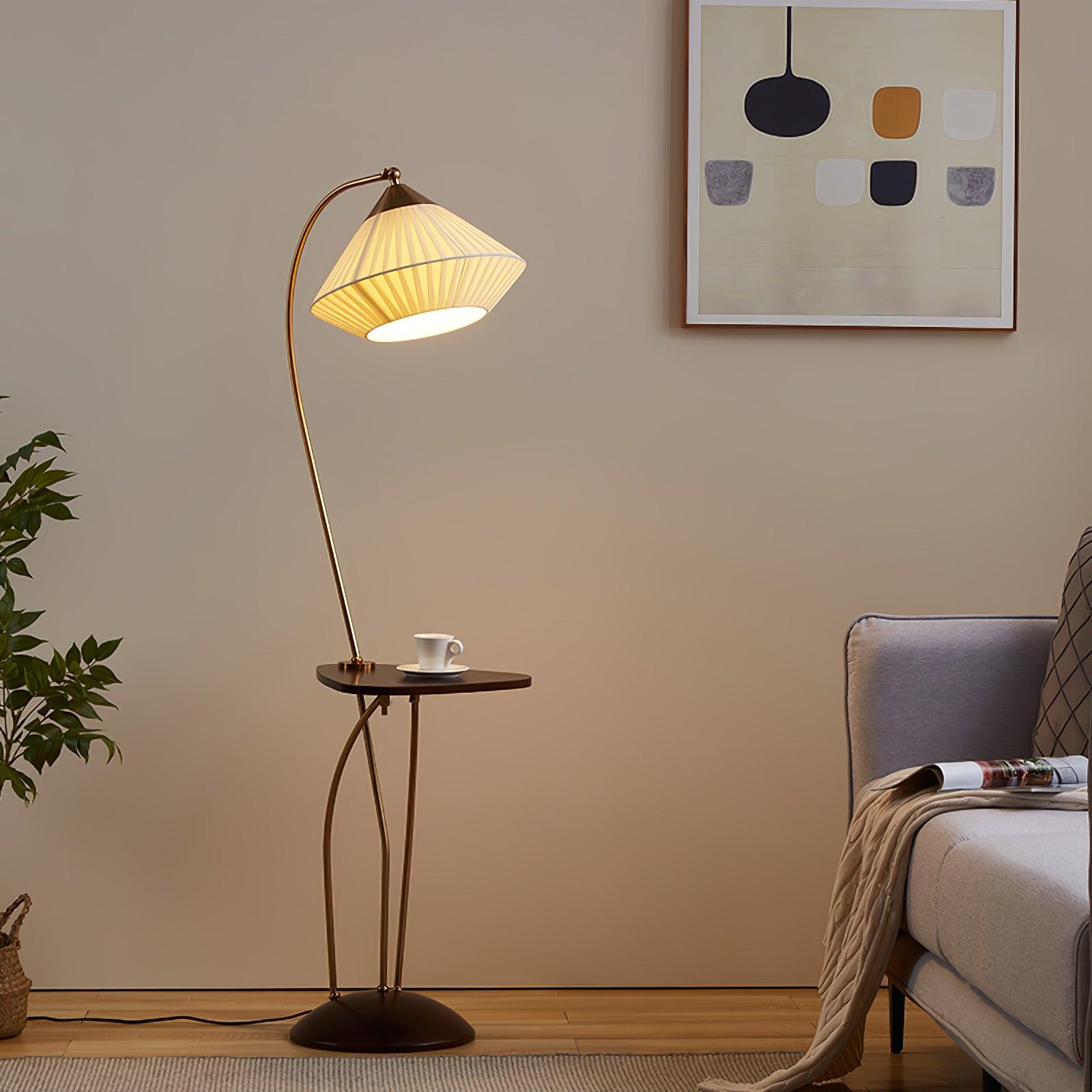 Curve lamp with a glowing pleated shade and cup on its small table beside a grey armchair, plant and framed art