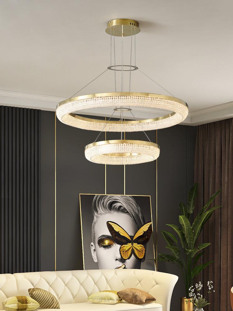 Two-ring gold crystal chandelier above a pale tufted sofa beside butterfly artwork and a tall plant