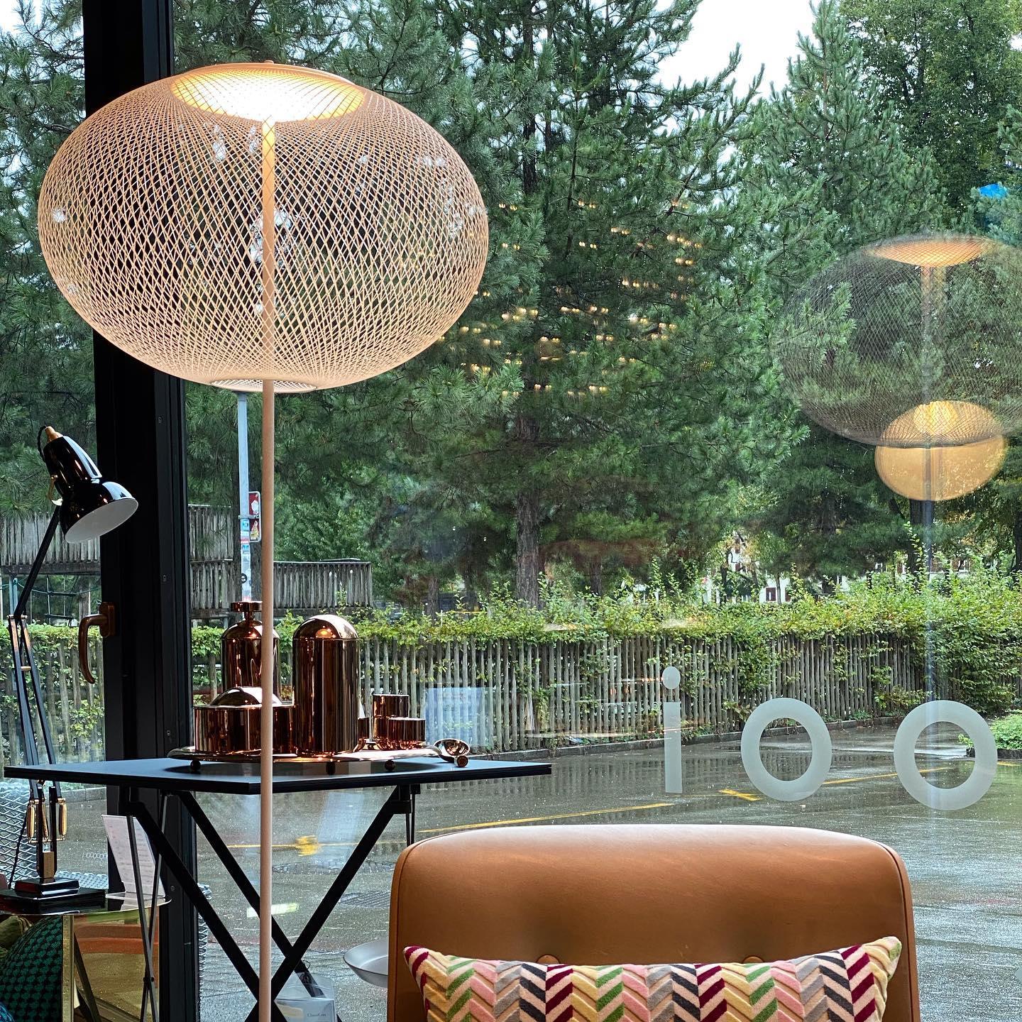 White mesh floor lamp glowing beside a tan chair at a large window, with trees outside and furnishings reflected in the glass