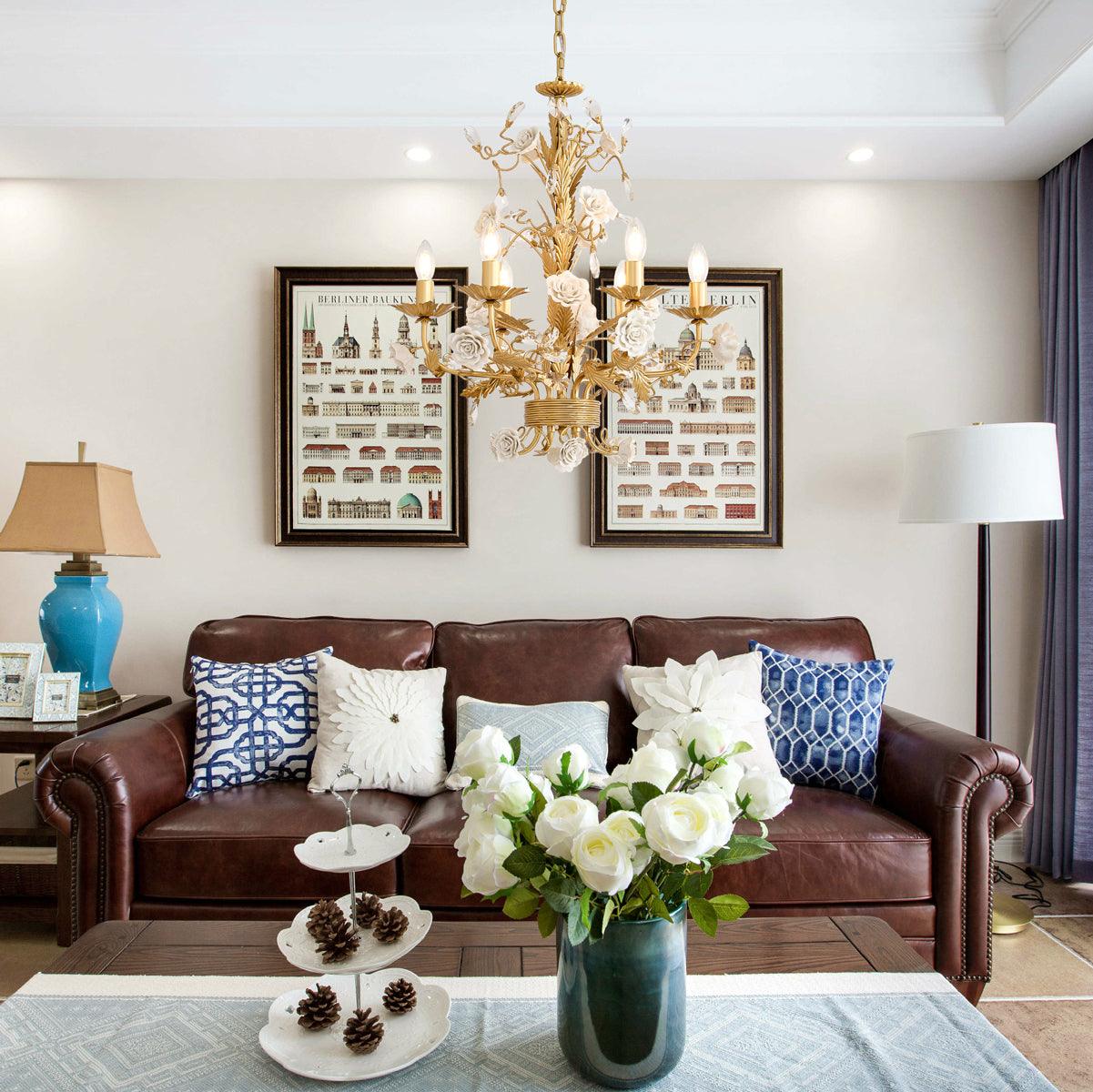 Lit floral chandelier over brown leather sofa with blue-and-white cushions, framed prints, rose vase and separate room lamps