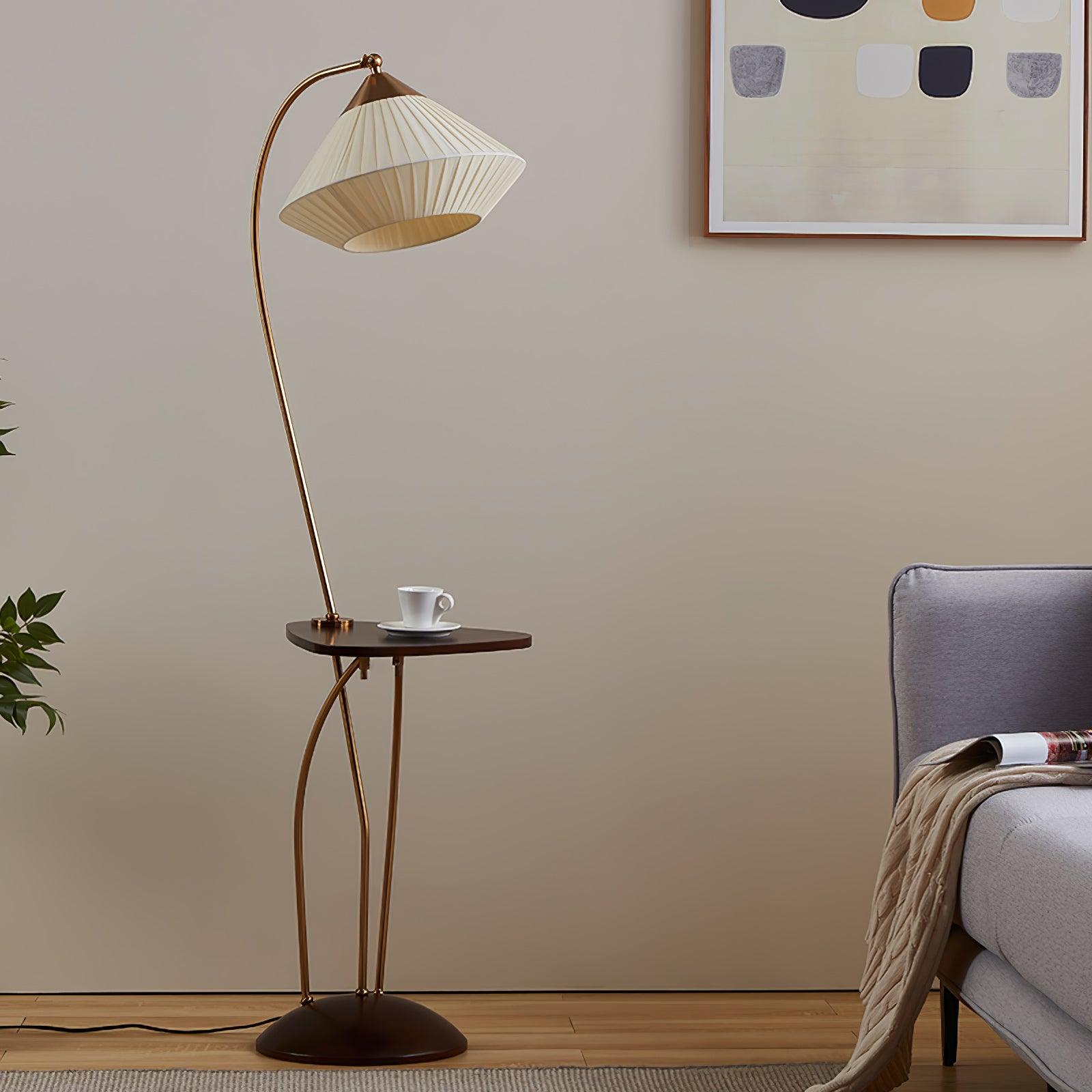Unlit Curve lamp beside a grey armchair with a magazine and throw, showing its beige pleated shade and dark table