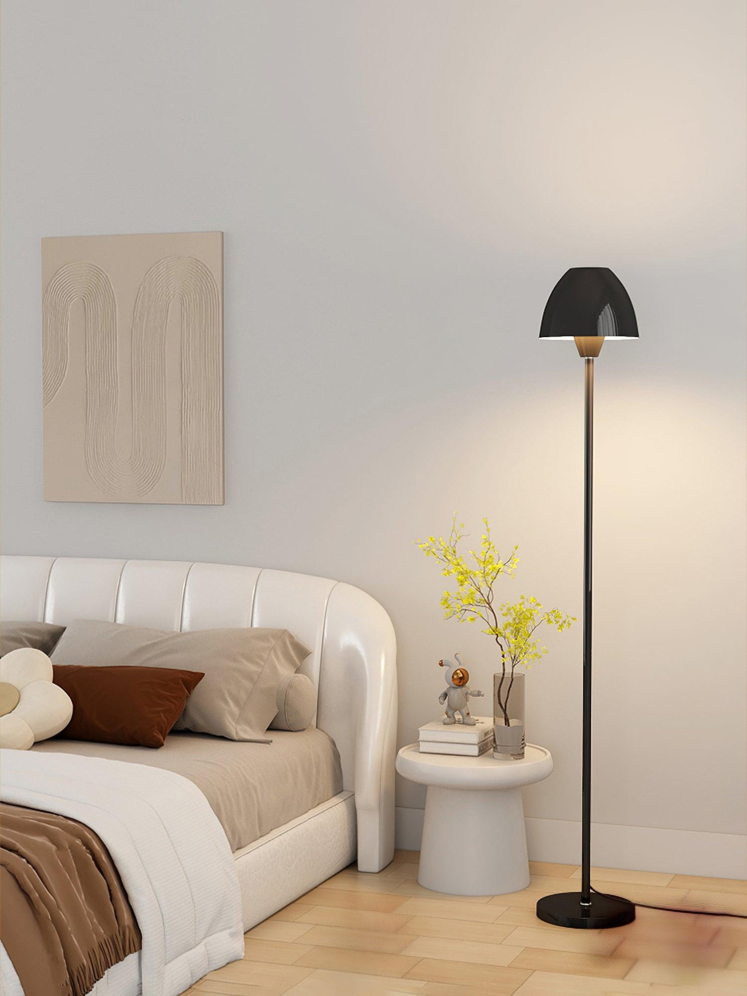 Black floor lamp beside a bed with a pale curved headboard and a round bedside table holding books and flowers