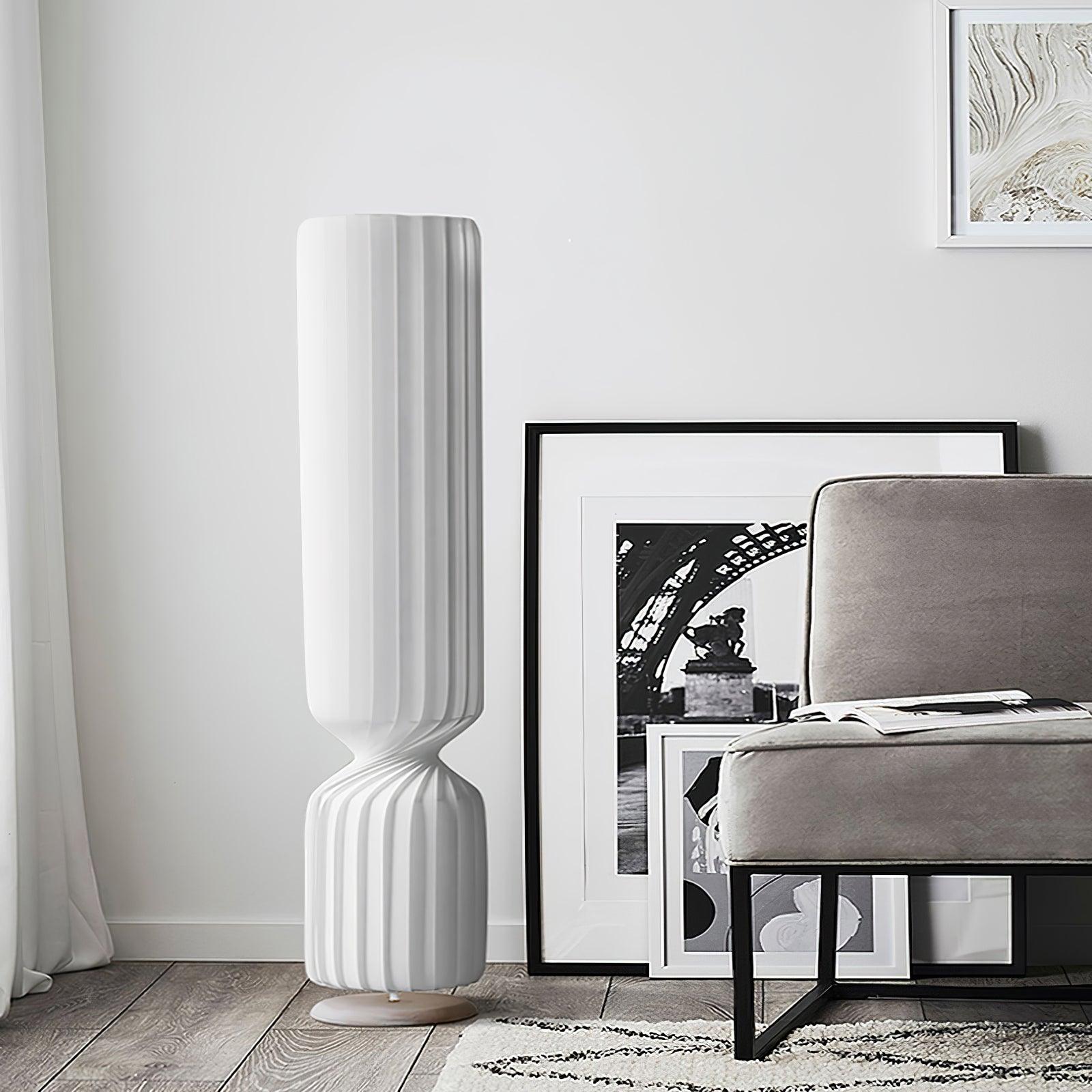 Close white pleated twist lamp beside gray chair and leaning framed prints, narrow waist and pale round base on wood floor