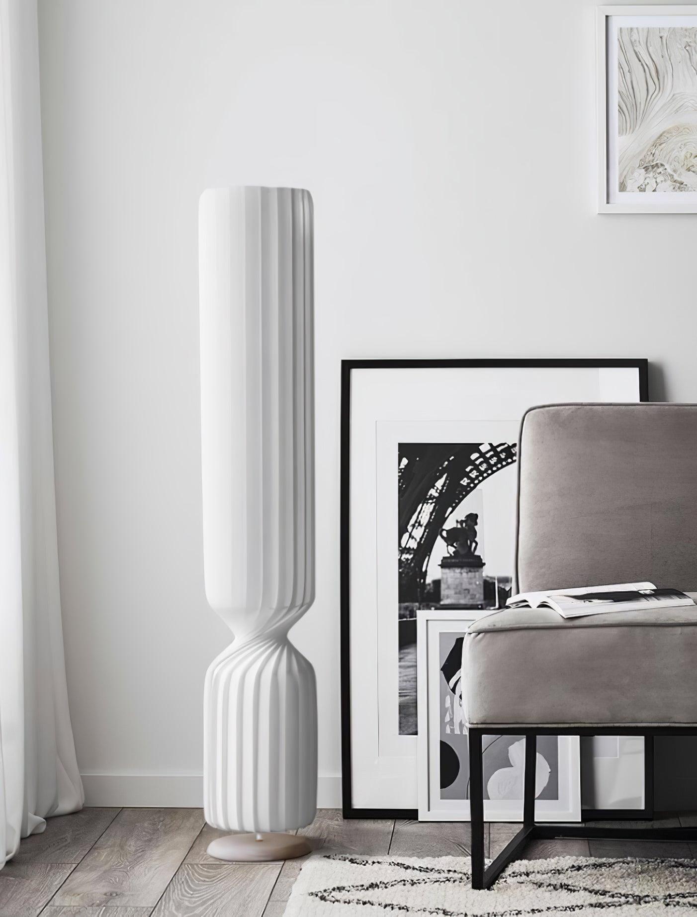 White twist lamp beside gray chair with open book and leaning monochrome prints, white curtain, wood floor and patterned rug
