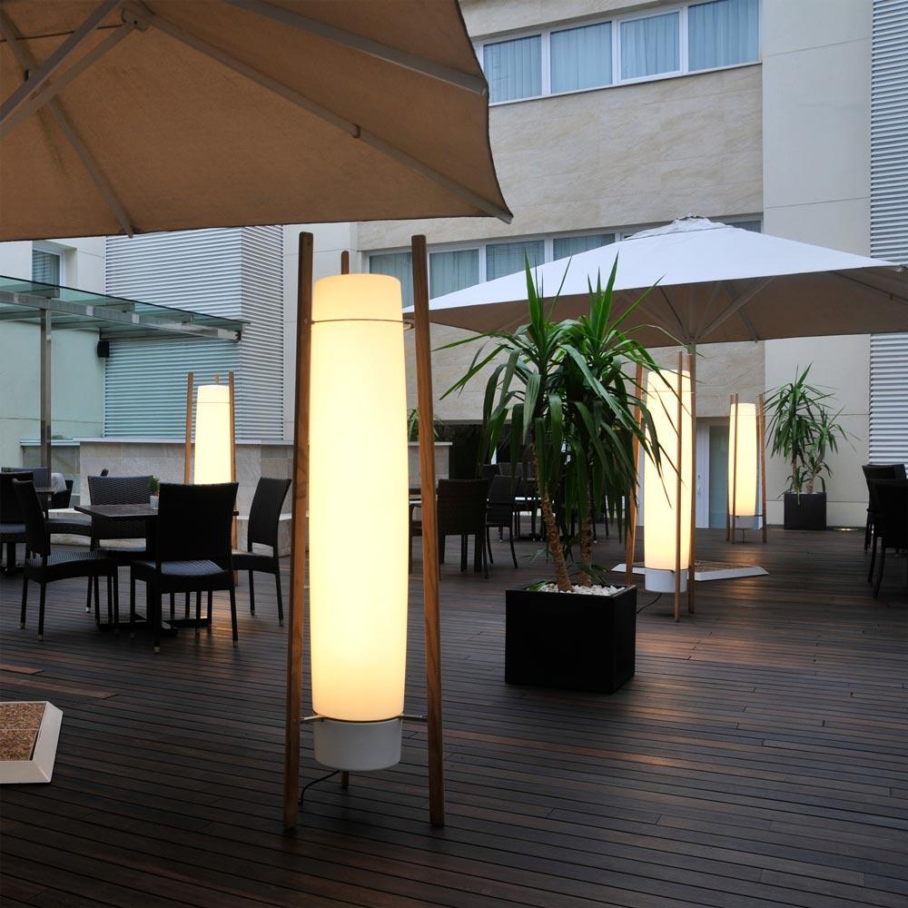 Several lit column lamps with wooden supports and grey bottom caps on a dining deck beneath parasols beside planters