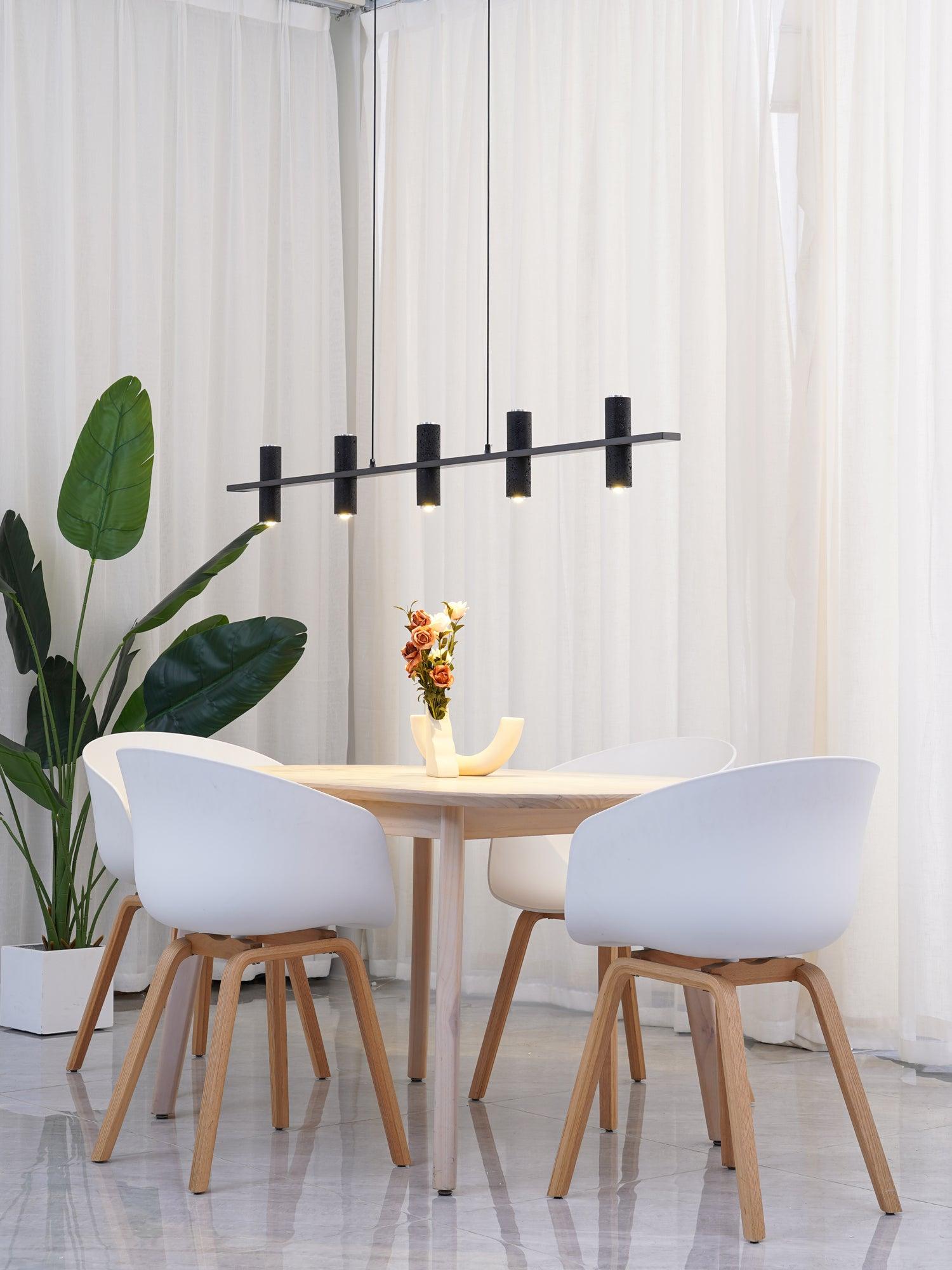 Side view five-cylinder chandelier over pale dining table and white shell chairs, with green plant in square pot and curtains.