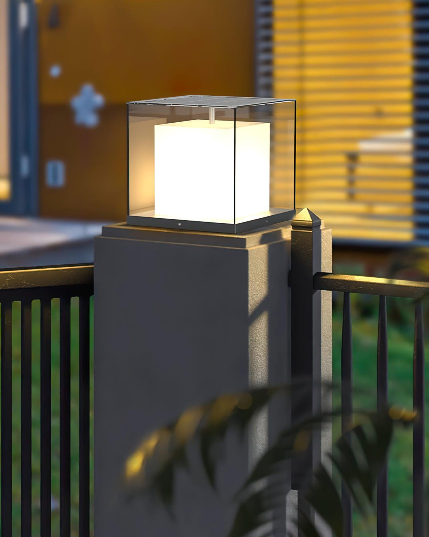 Single glowing glass cube light on a grey gate pillar beside a dark rail fence and shiny finial, with foliage behind