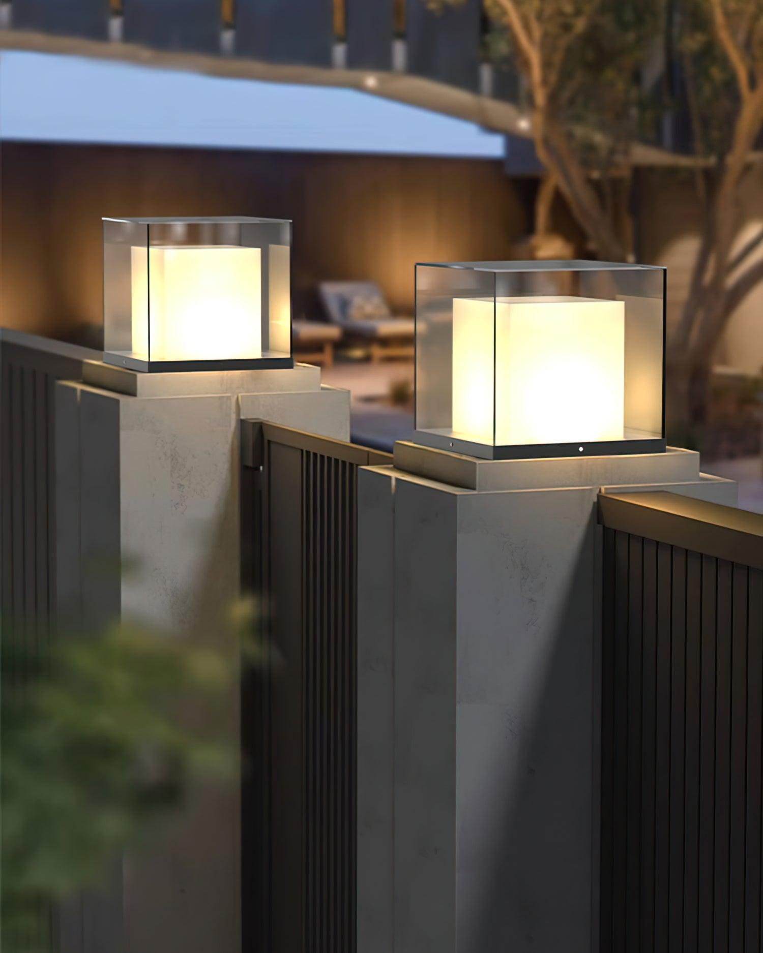 Two illuminated glass box lights on dark caps of pale gate pillars above a black vertical-slat gate