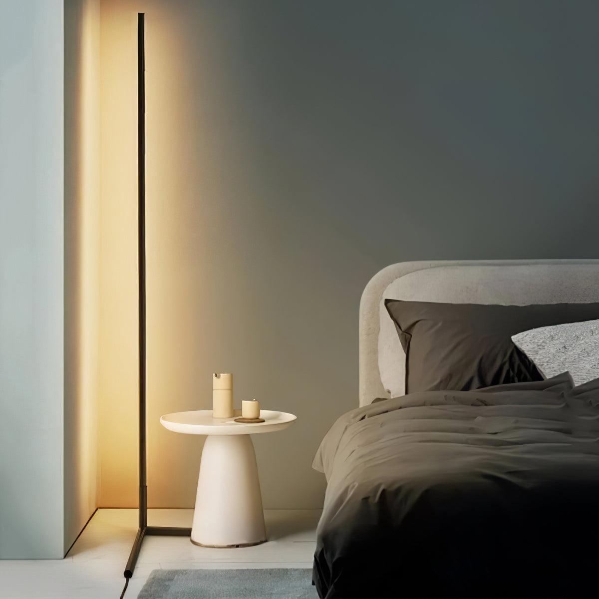 Warm-lit corner lamp with angled feet beside an upholstered bed and white cylindrical bedside table