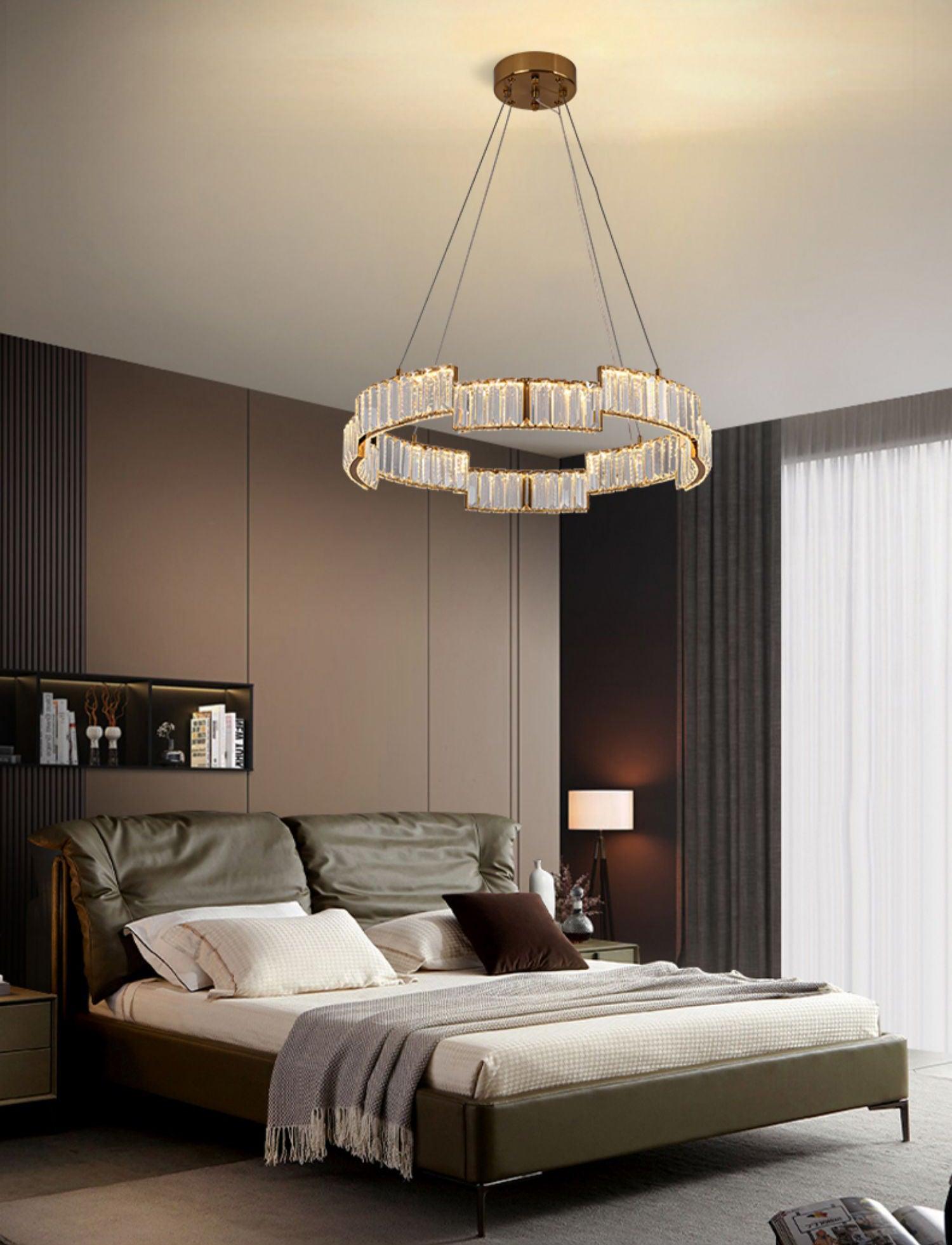 Lit glass ring chandelier above a green upholstered bed with pillows, brown wall, shelving and a separate lamp