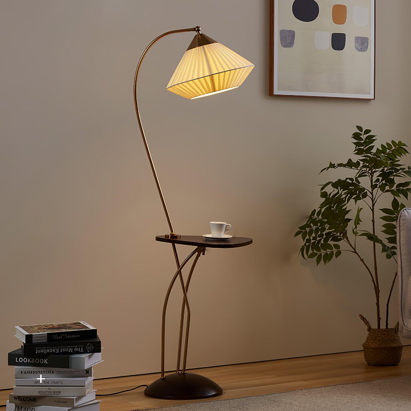 Curve lamp with a glowing pleated shade and cup on its dark table beside stacked books, a plant and grey chair
