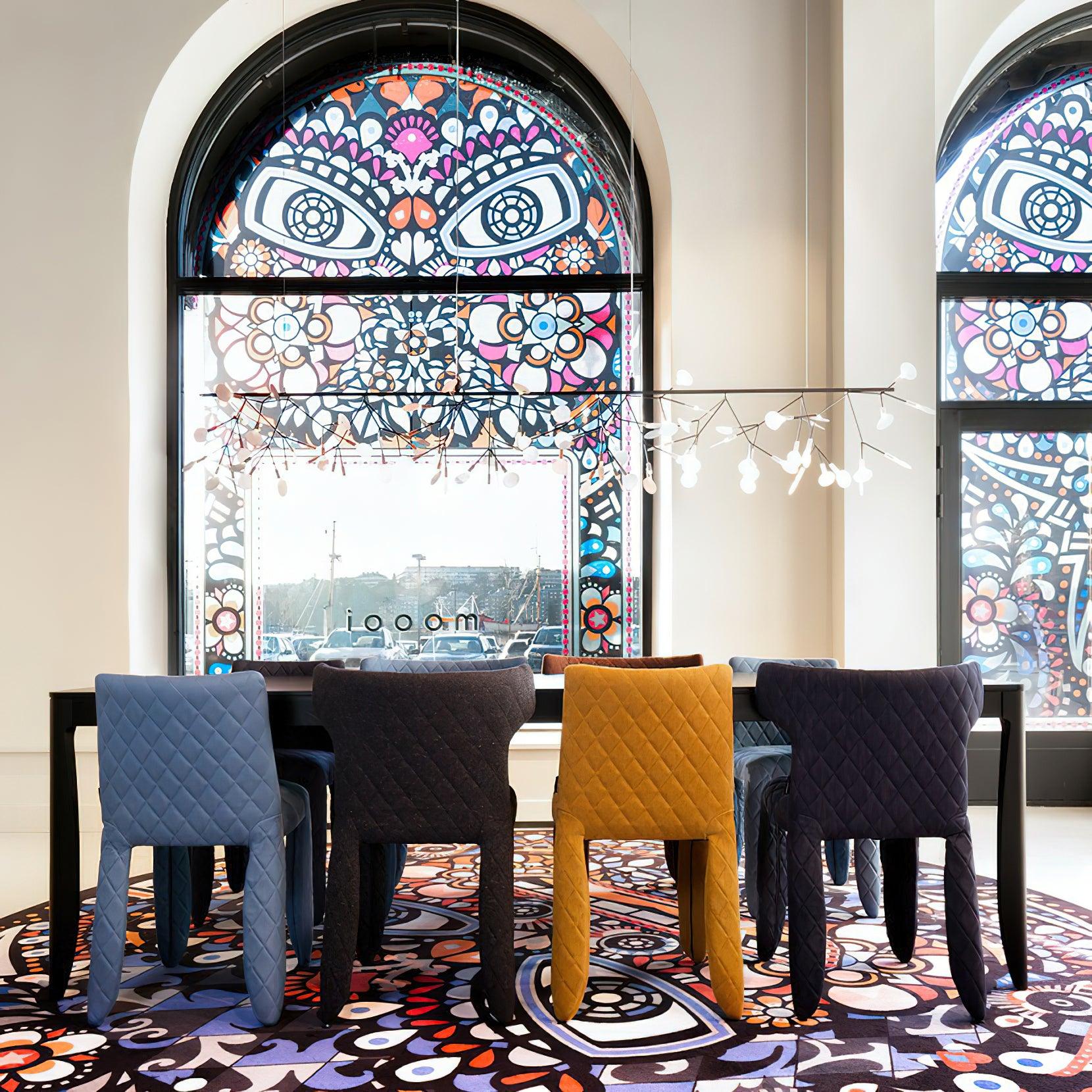 Linear petal chandelier above black table and quilted blue, grey and mustard chairs beside patterned arched windows