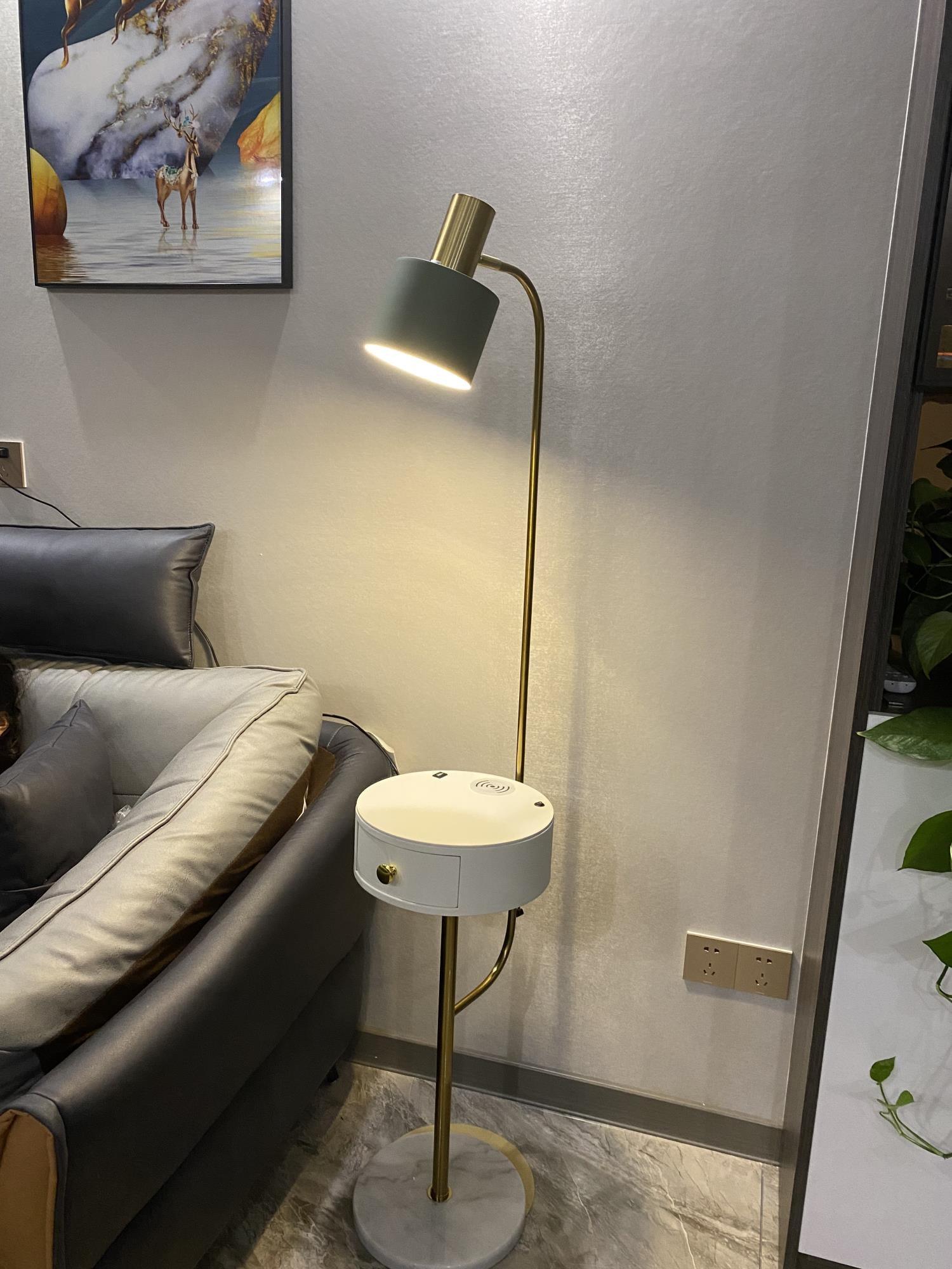 Gold lamp lit beside gray sofa, green shade, white closed-drawer table with small symbols and openings, white veined base