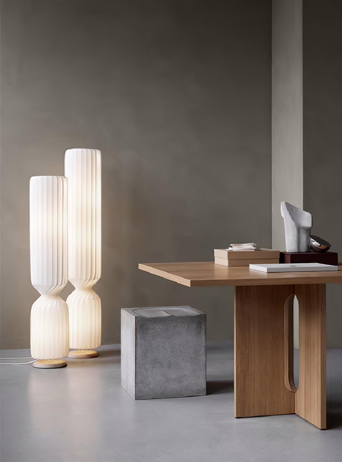 Two lit white twist lamps by wooden console with books and sculpture, gray block stool in front and cords across floor