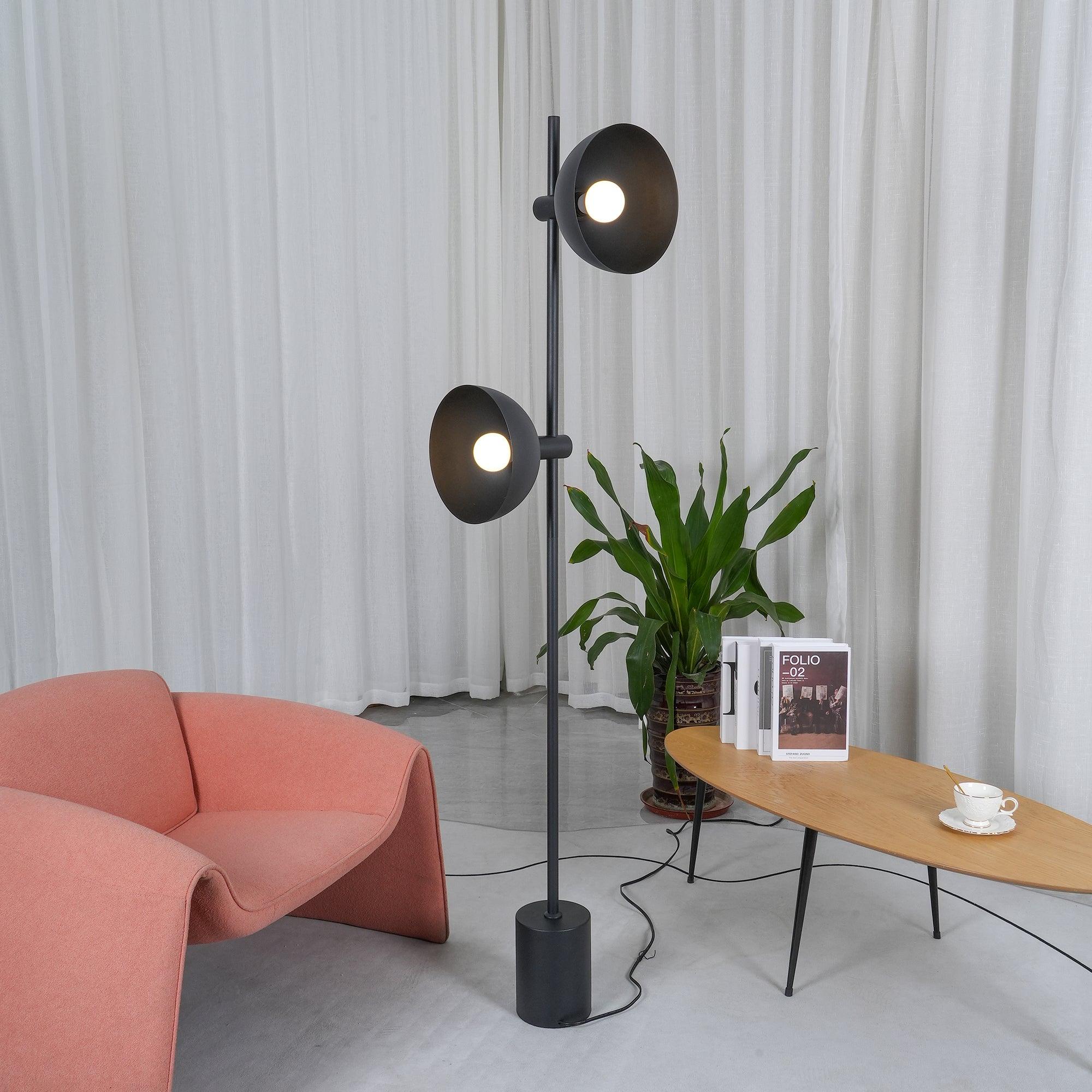 Studio floor lamp with both heads lit beside a coral armchair and wooden table.