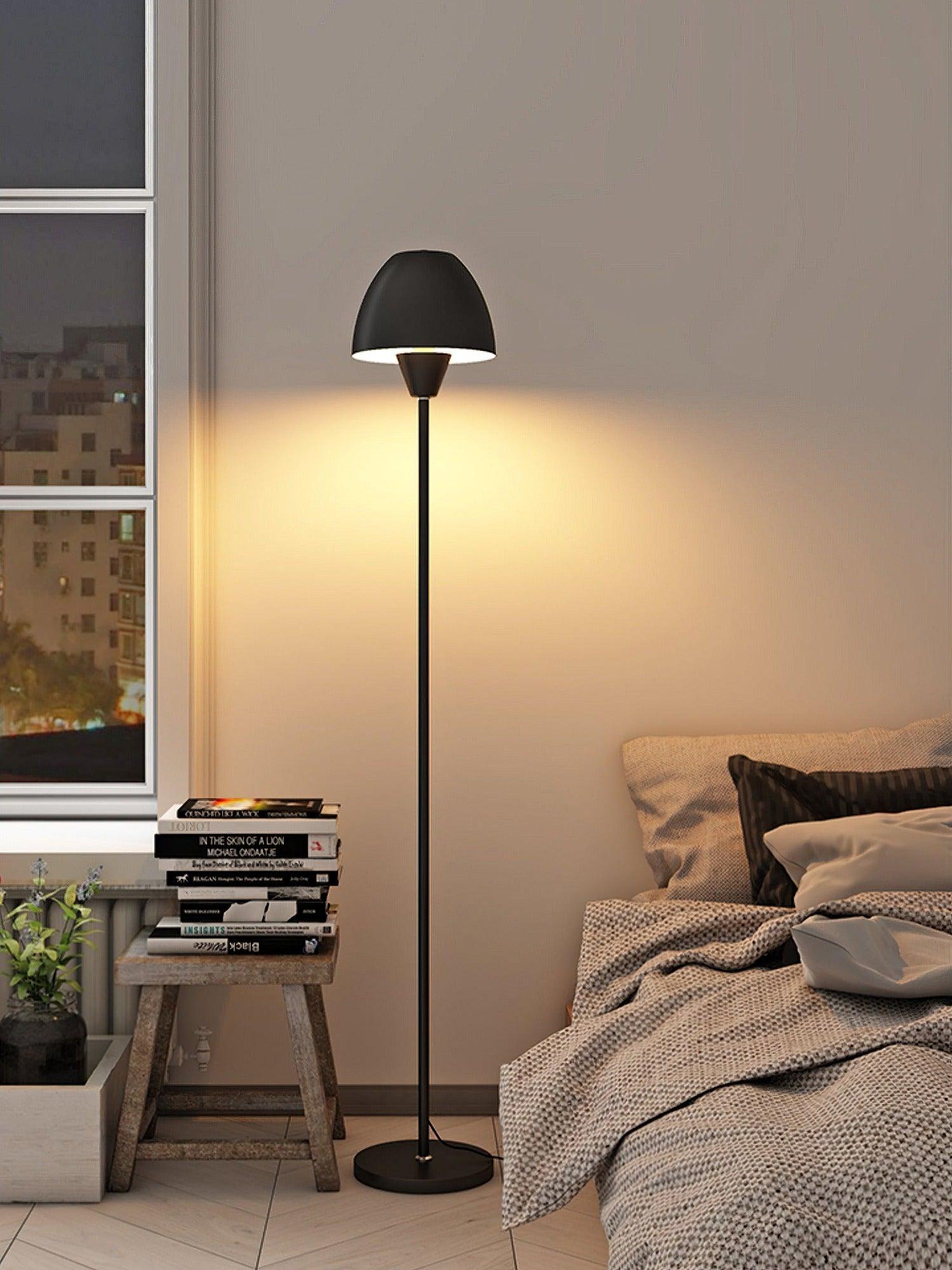 Black floor lamp by a bedroom window beside a bed and a wooden stool stacked with books
