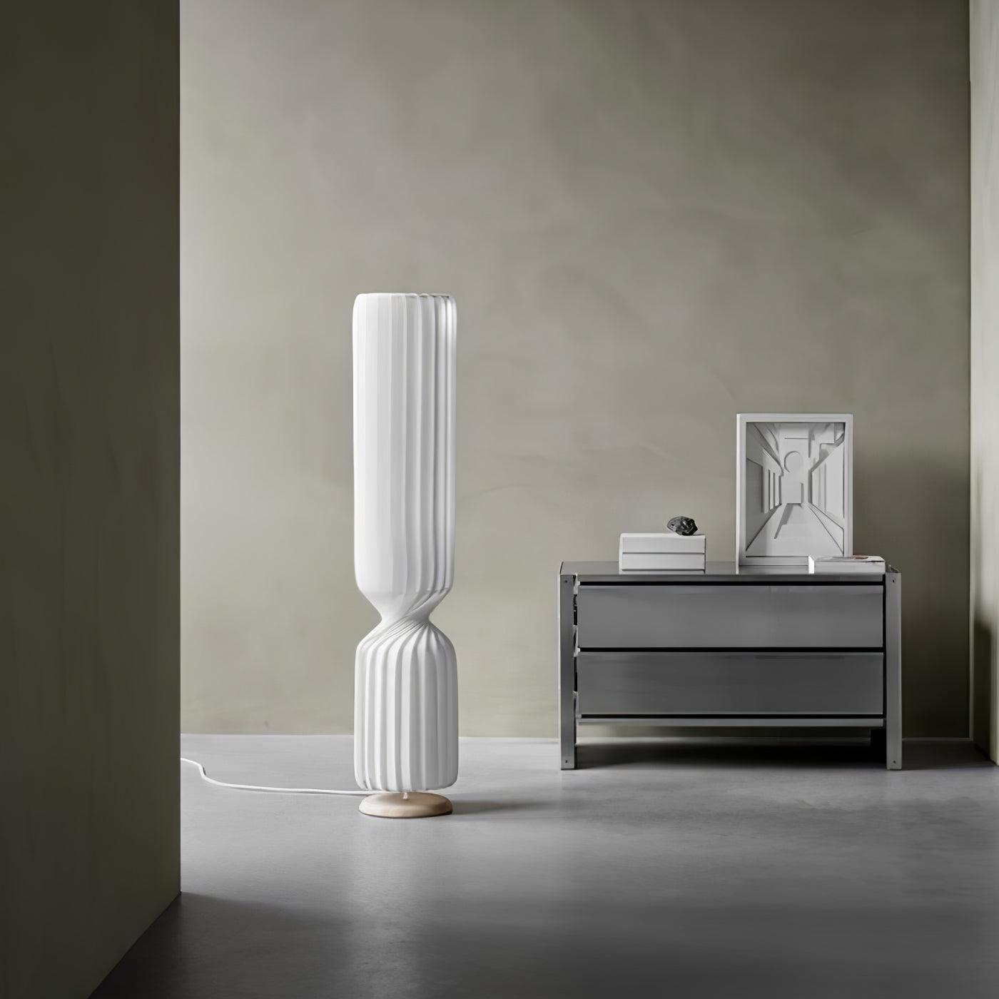 White twist floor lamp beside gray chest with framed perspective print and books, pale round base and white cord