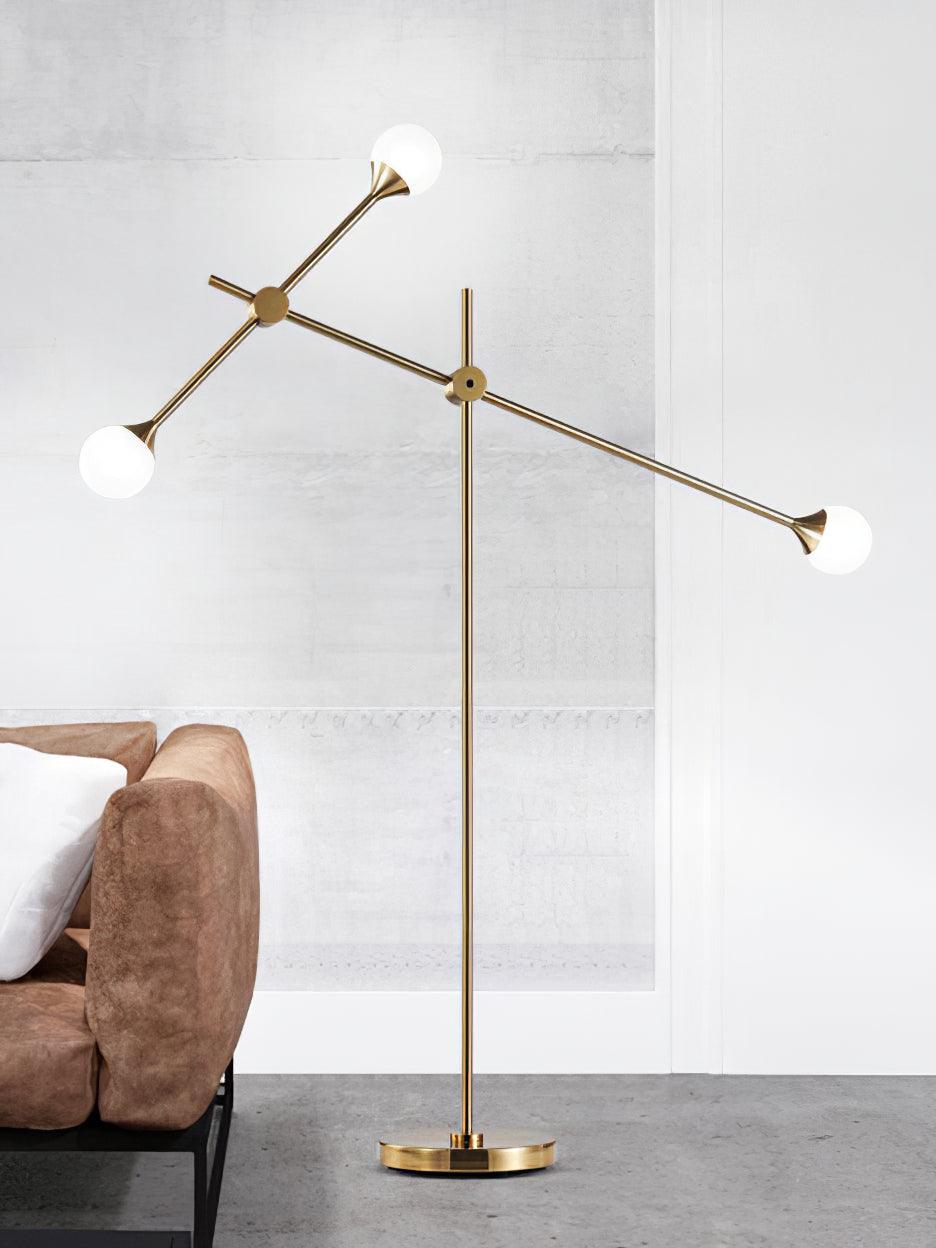 Full view of gold Kemi floor lamp with three white globes beside a brown sofa on a grey floor