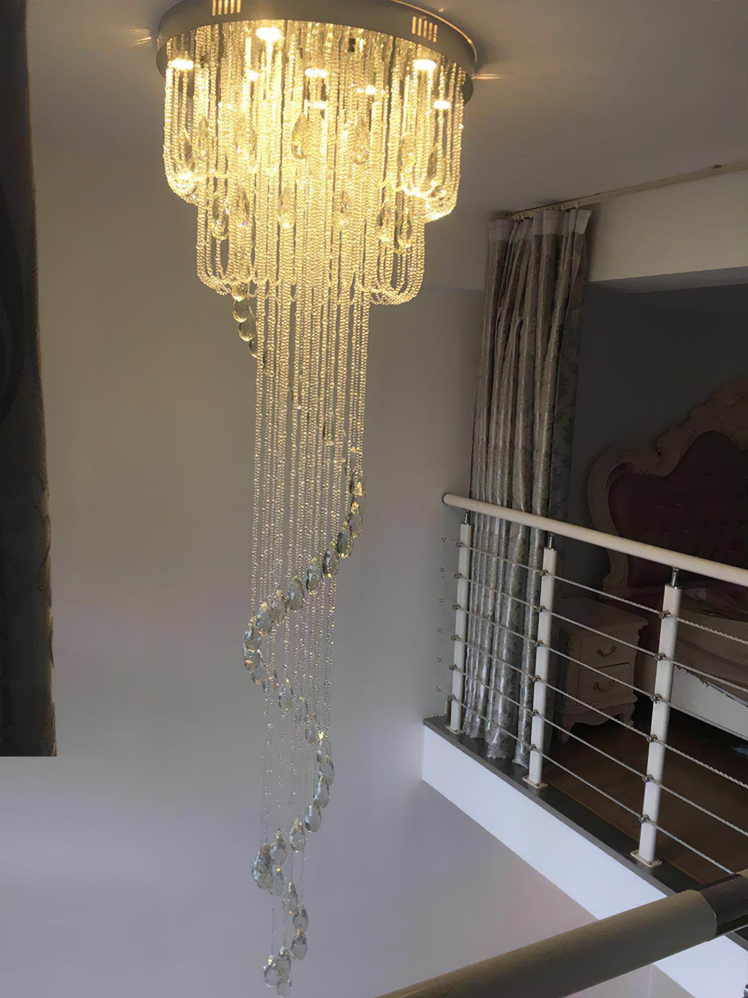 Warm-lit crystal spiral beside an upper-floor landing with horizontal cable railing, curtains and dark upholstered furniture